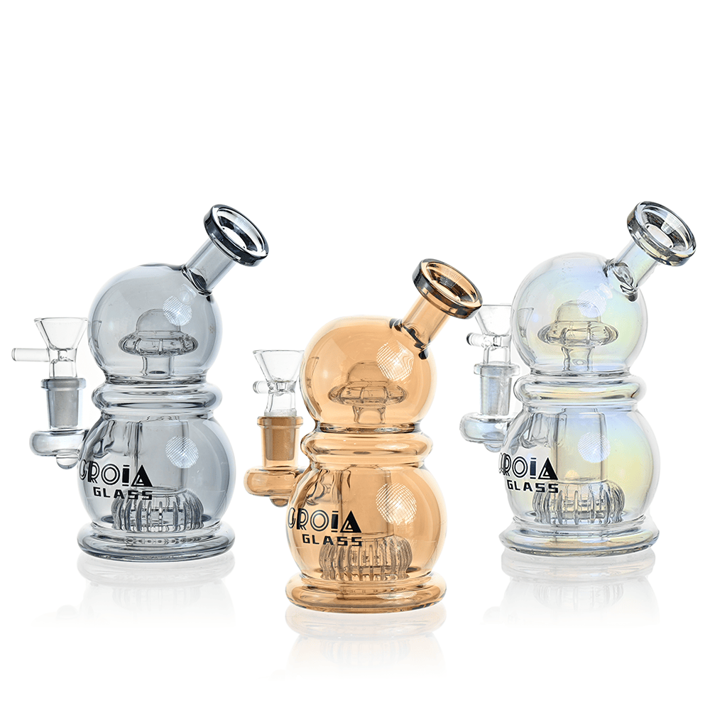 Three-quarter front view of three Croia Glass 6" Mini Egg Dab Rig with Crown Perc pieces in navy-toned, amber, and iridescent glass arranged side-by-side on a white background