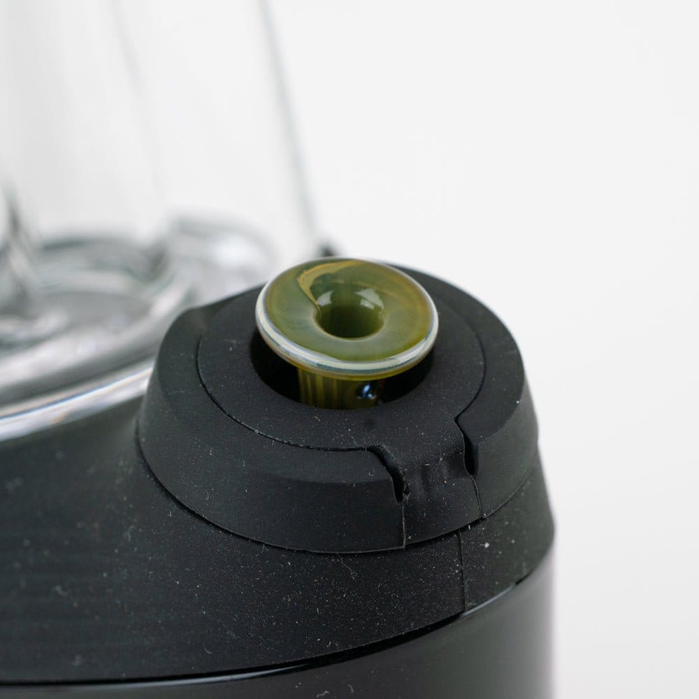 Close-up angled shot of the Empire Glassworks PuffCo 'New Peak' & Proxy Glass Joystick Cap - Vintage sitting in a black rubberized mount, showing the olive-green glass cap detail and nearby clear glass chamber.