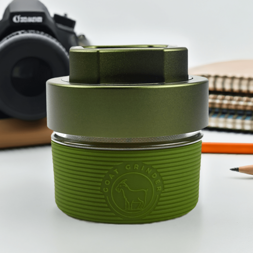 Front view of the AITH v.I – OG Grinder Jar, with a smooth, rounded form displayed beside a camera and patterned cloth.