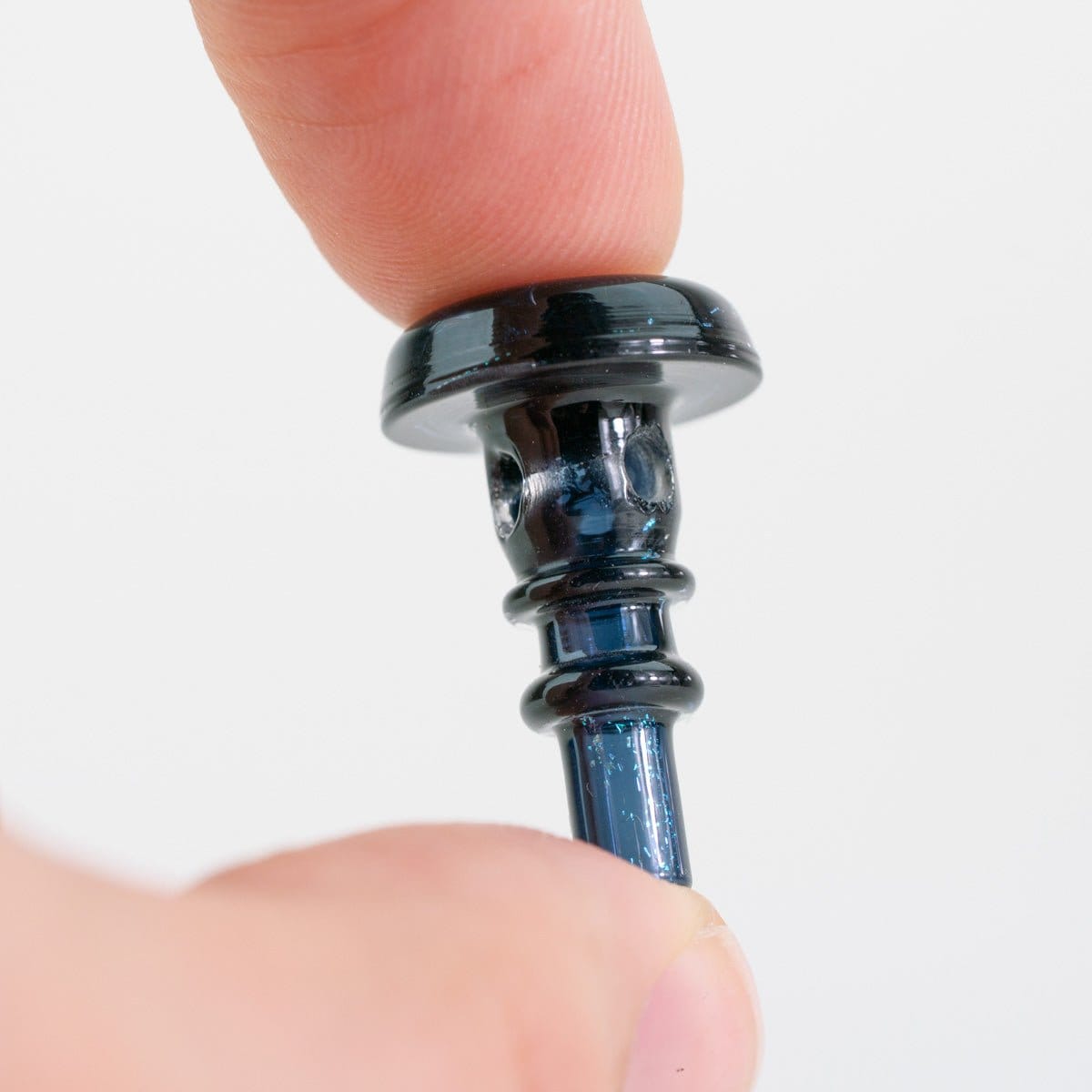 Close-up of a black, iridescent Empire Glassworks PuffCo Peak Pro 3D XL Chamber Glass Joystick Cap - Black Nebula held between thumb and forefinger against a plain white background
