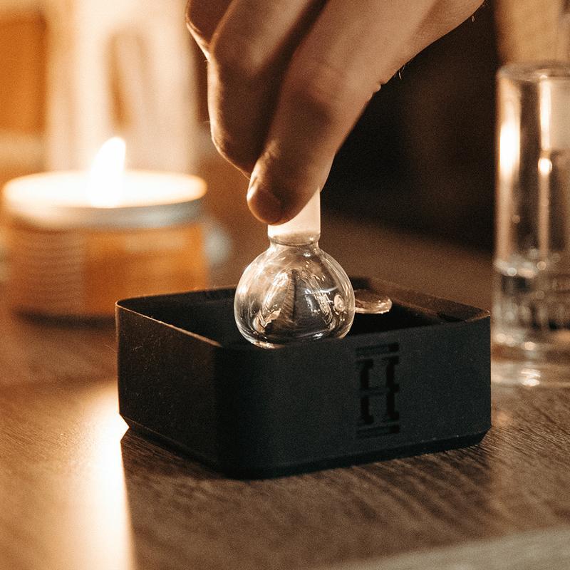 A dimly lit close-up showcases the Silicone Caché Debowling Ashtray resting beside a lit candle and glass, emphasizing its sleek, modern design.