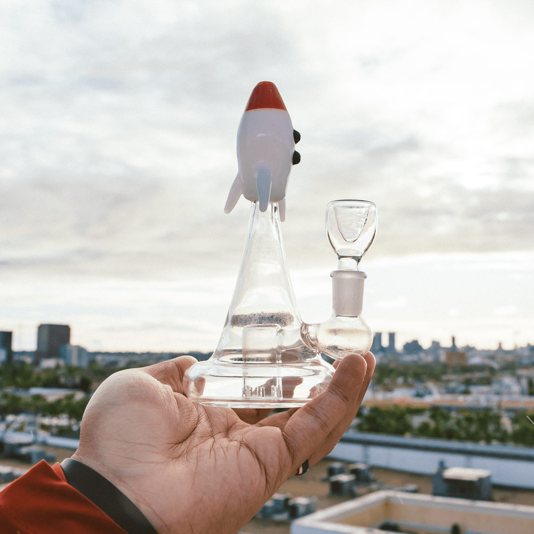 A hand holds the Blastoff Rocket Bong against a blurred city background, showcasing its unique rocket-shaped design.