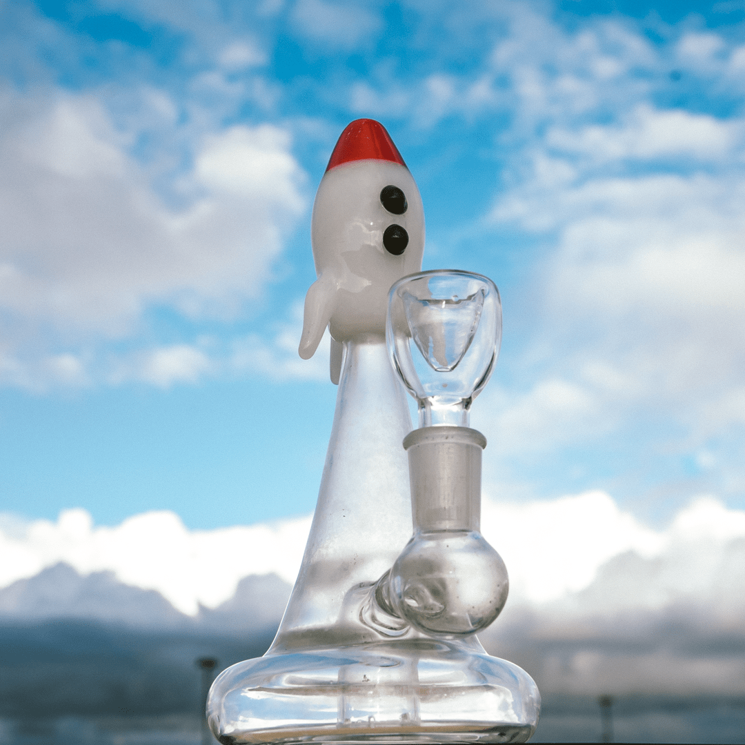 Front view of the Blastoff Rocket Bong, a clear glass water pipe shaped like a red-tipped rocket ship against a blurred blue background.