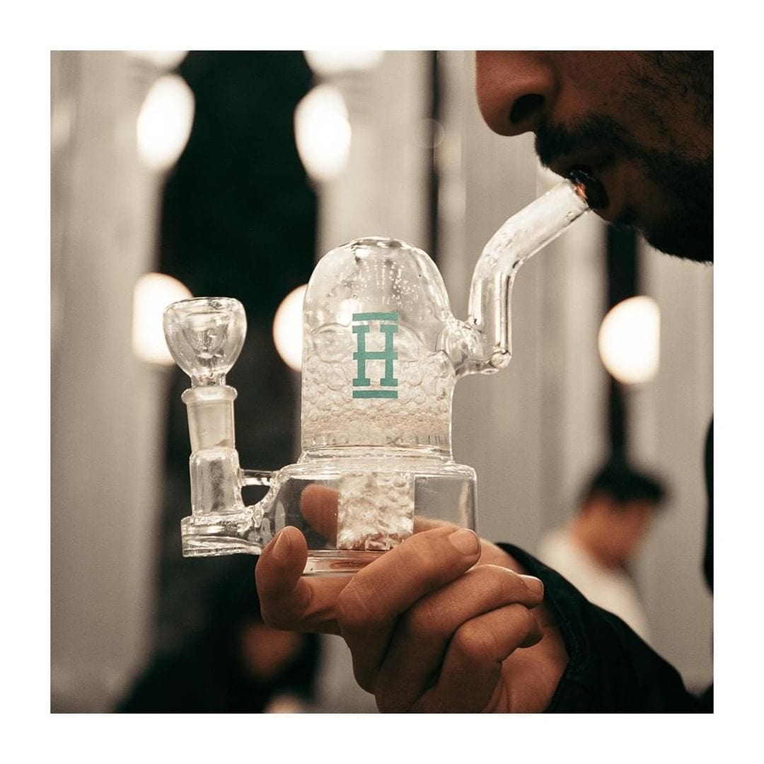 Close-up side view of a person using the Hemper Bell Rig, a clear glass bubbler with a domed chamber and teal "H" logo.