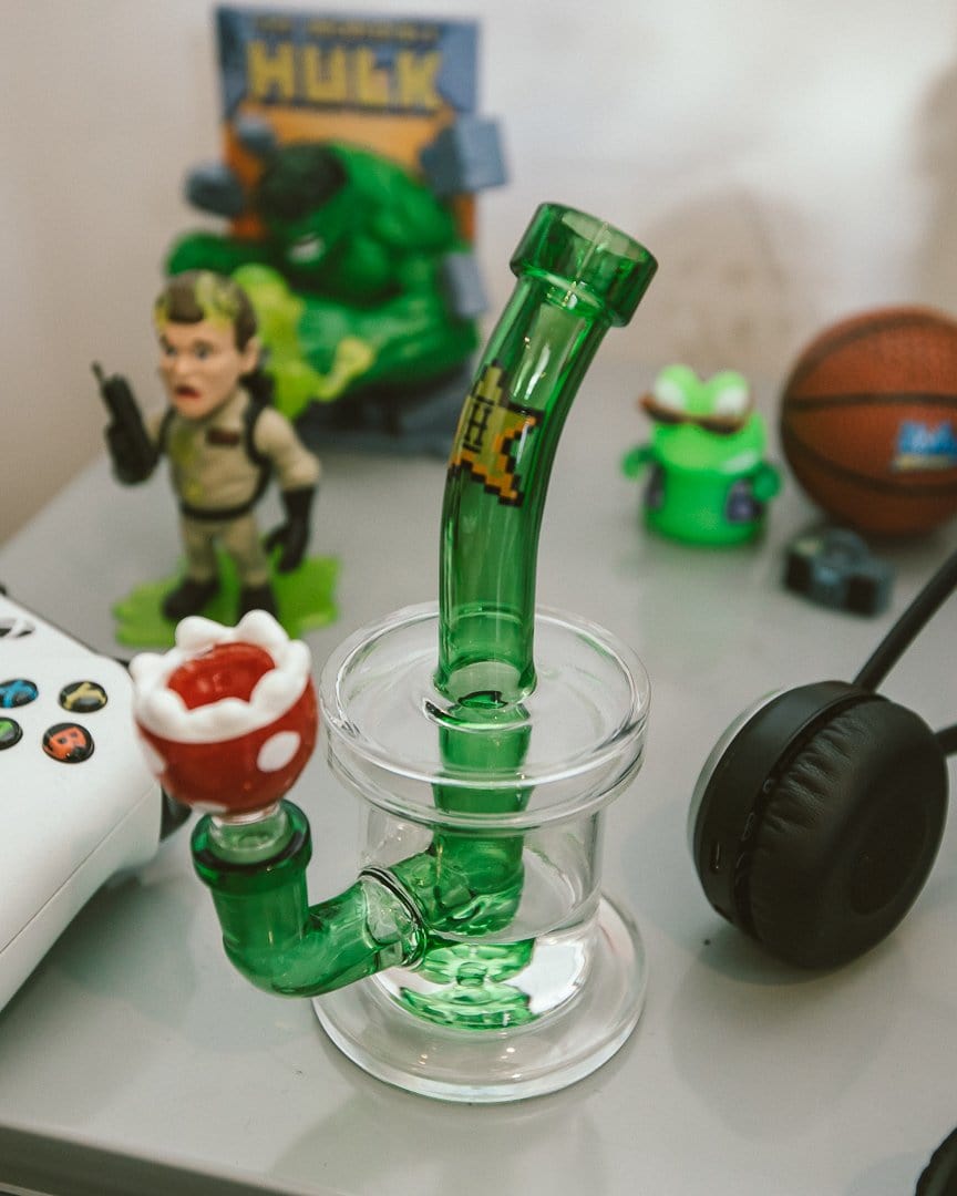 A front view of the Hemper Gaming Bong amid gaming accessories and figurines on a light grey surface.