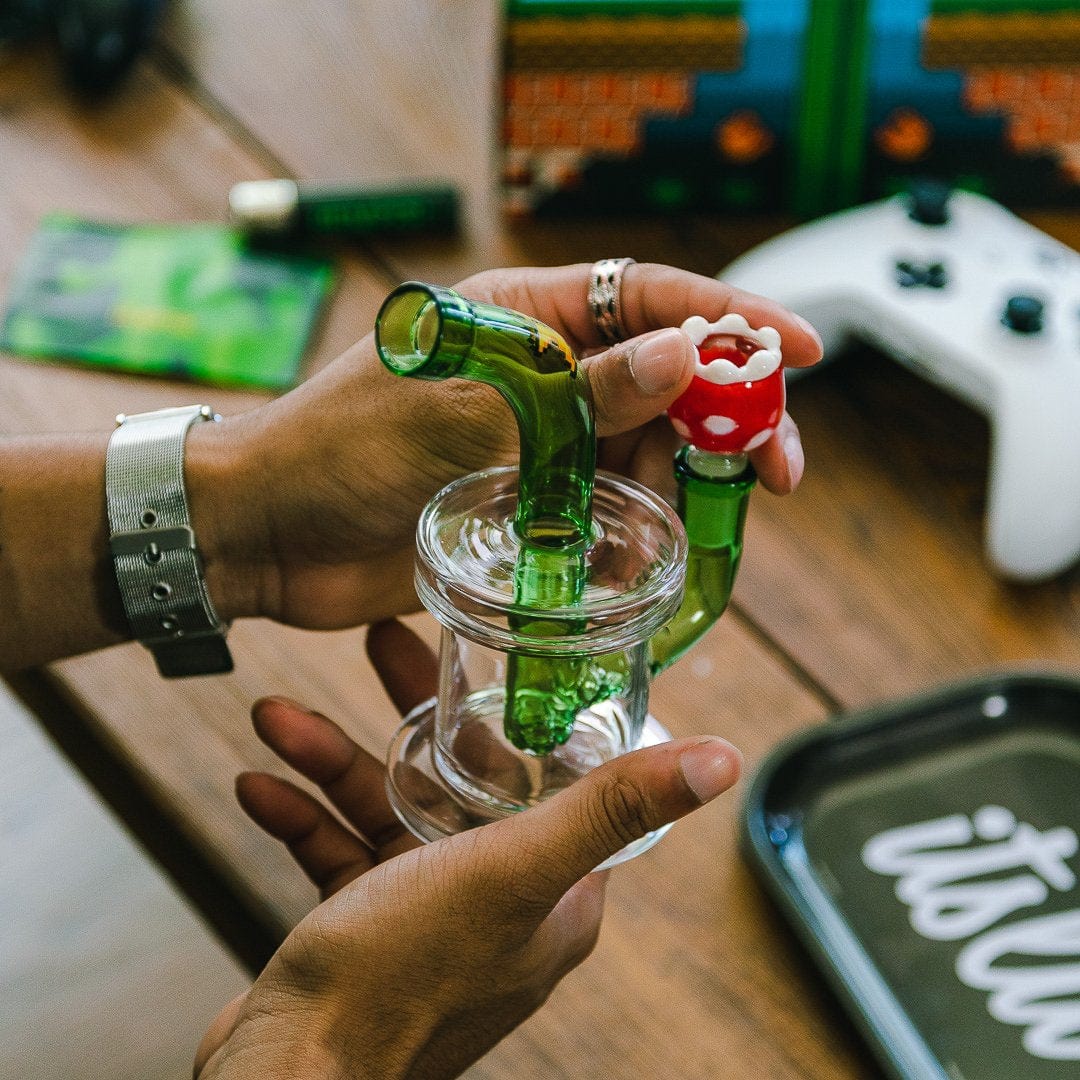 A close-up, angled view showcases the Hemper Gaming Bong held in a hand, alongside a white gaming controller.