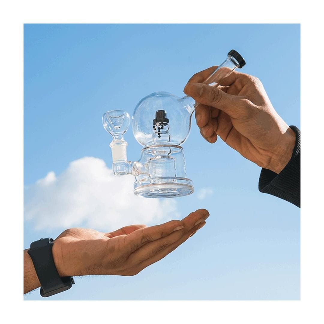 A pair of hands display the Snow Globe Bong against a bright blue sky, highlighting its clear glass construction.