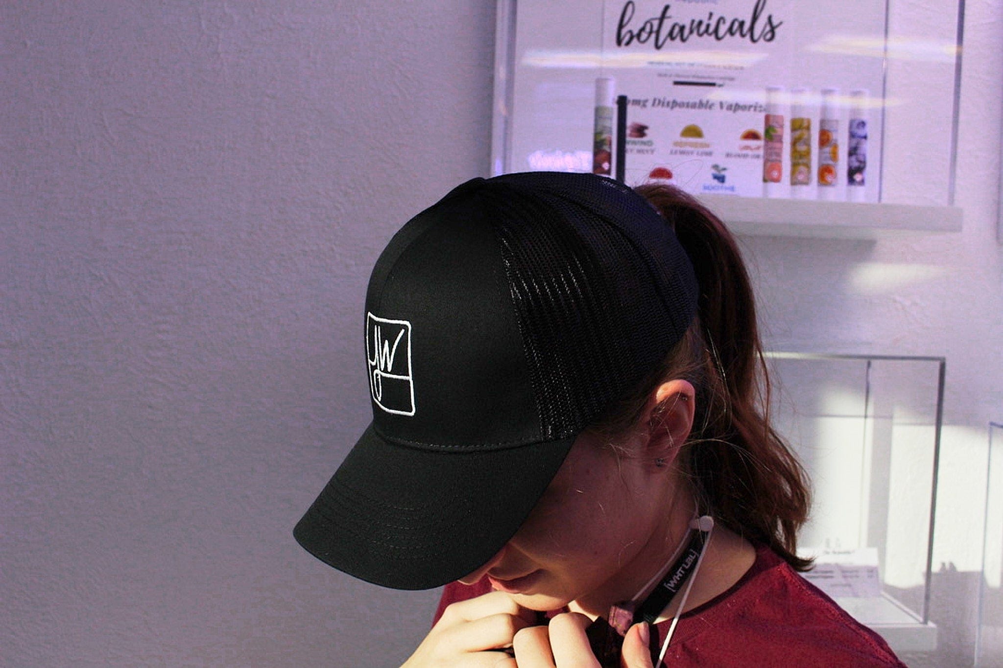 Top-down view of a person wearing a black Jane West Hat with a mesh back, head tilted down against a pale purple wall.