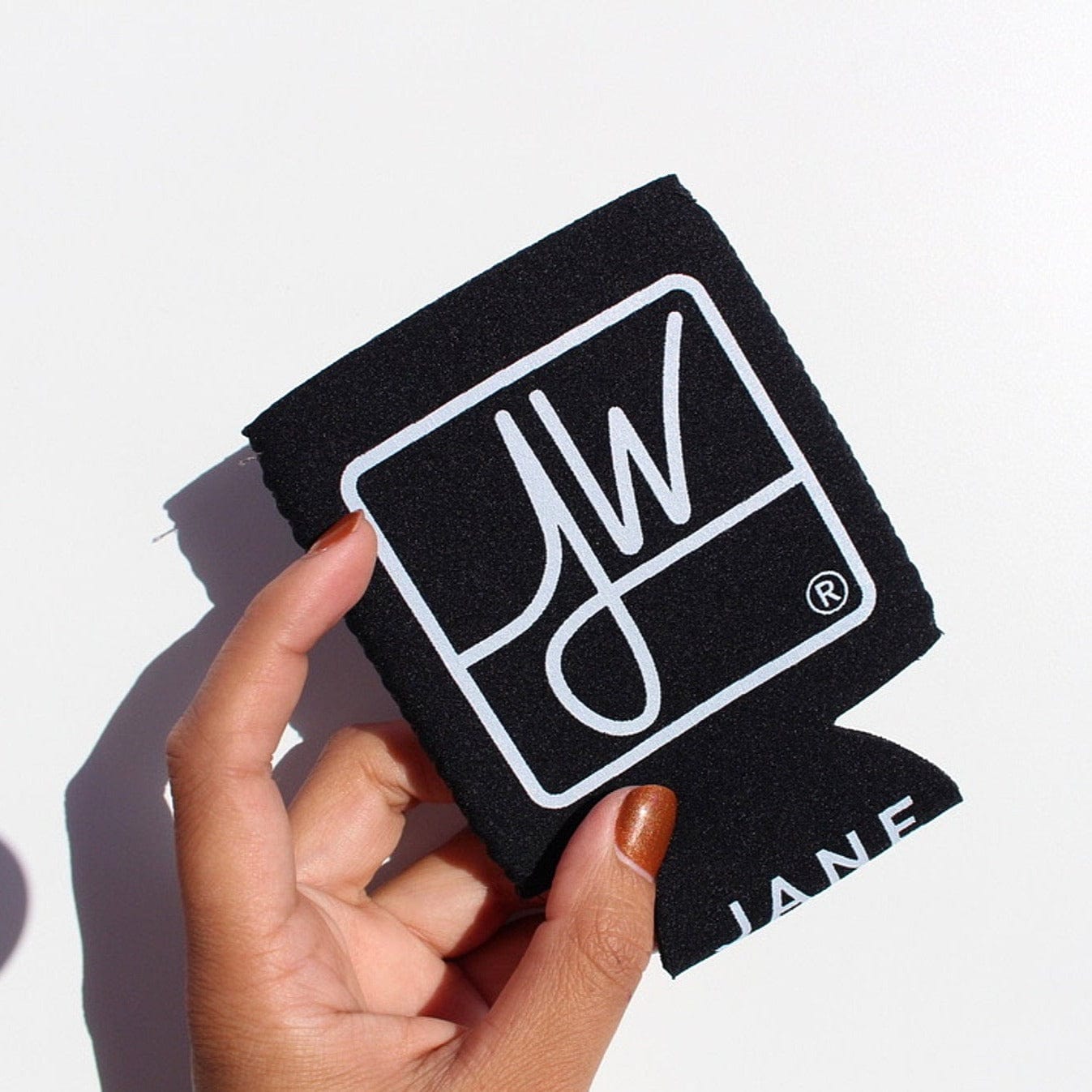 Handheld close-up of the black Jane West Koozie with Custom Pocket showing a white JW logo against a bright white background.