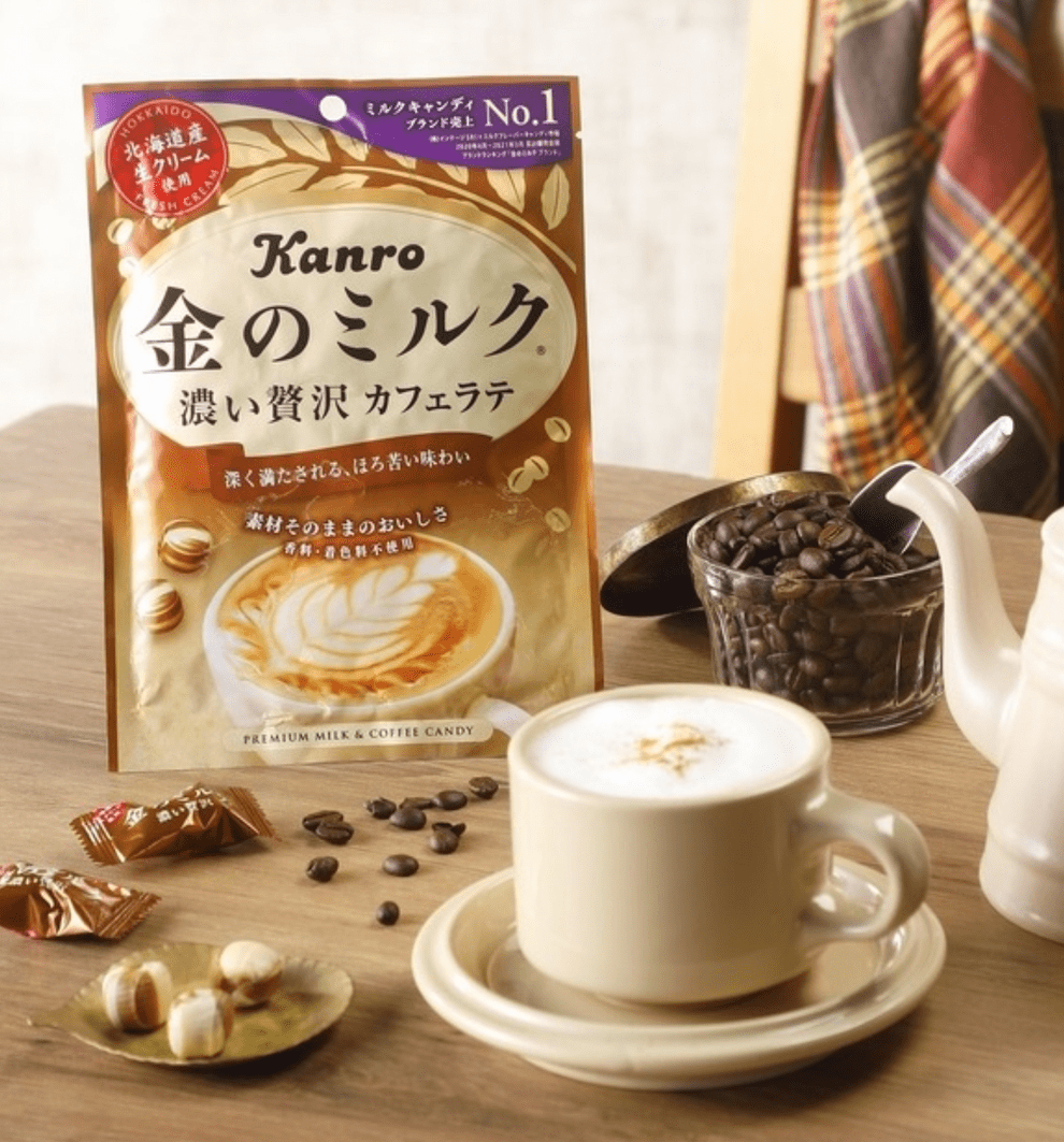 A close-up, angled shot features Kin No Milk Candy - CafÃ© Latte candies arranged beside a cup of coffee on a wooden surface.
