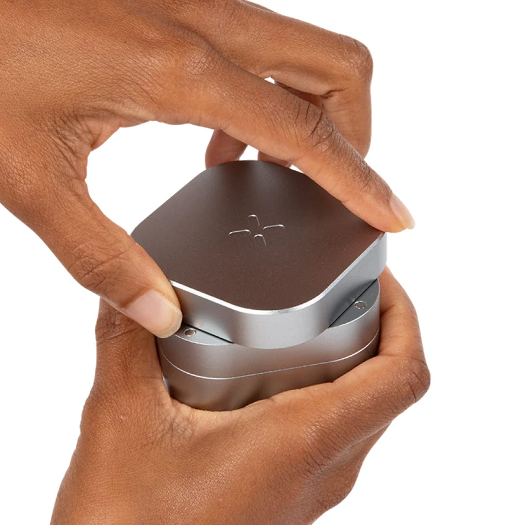 A close-up, angled view shows a hand holding the silver PAX Grinder, with a focus on its textured, circular top.