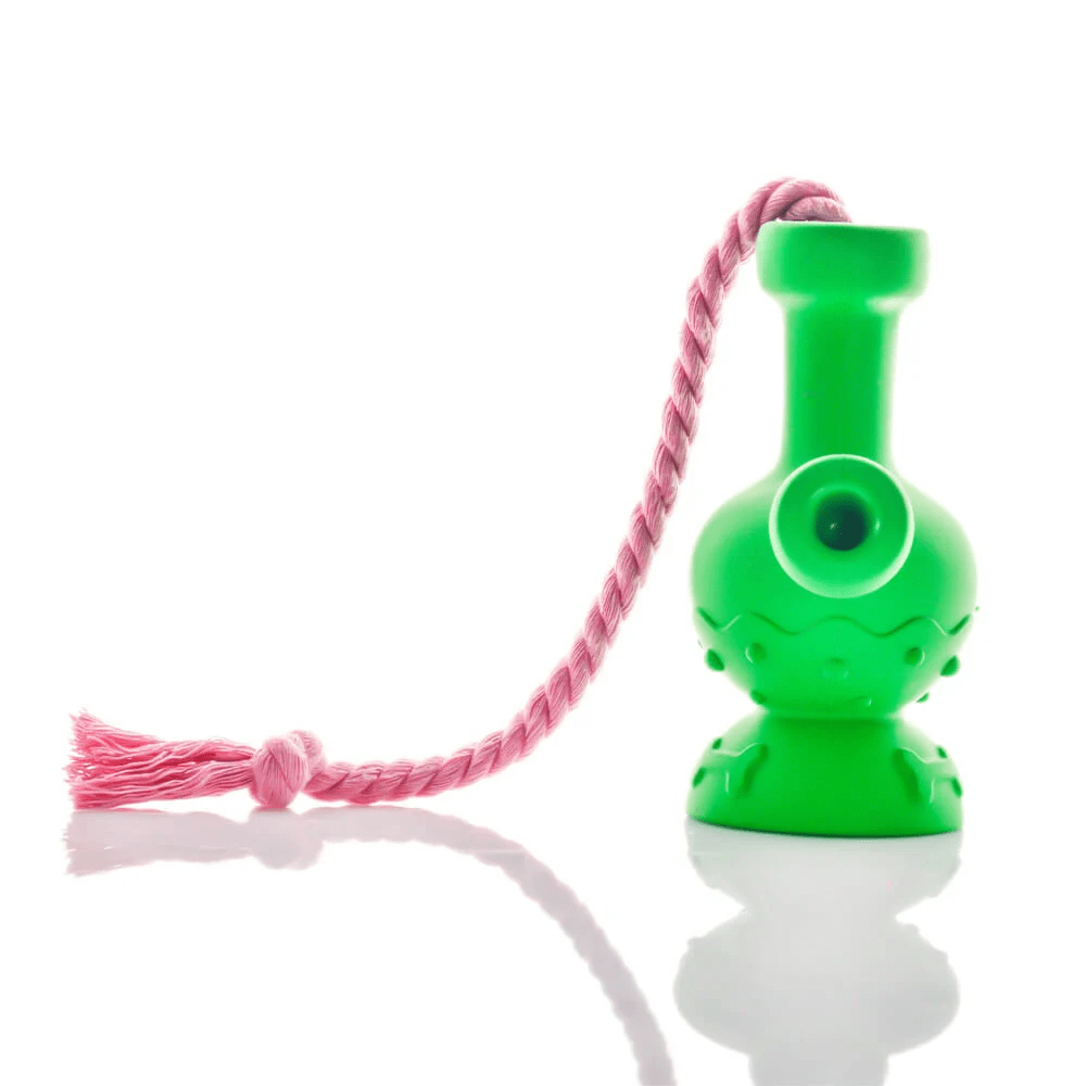 Studio-style side view of a bright green Tug & Toke with a coiled pink tassel draped beside it on a reflective white surface