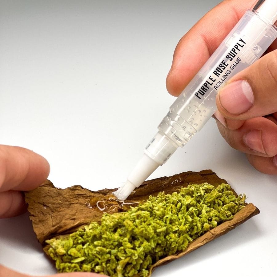 Close-up of hands using a Cannagar & Blunt Rolling Glue - 4ml pen to apply clear adhesive to a brown leaf filled with green herb.