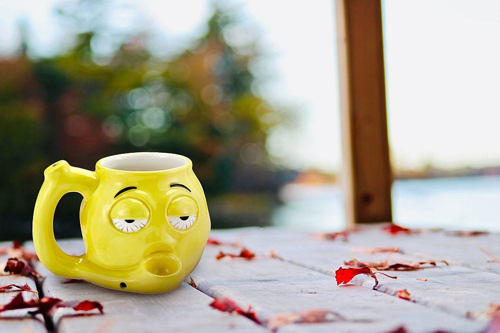 A front view of the yellow Stoned Emoji Pipe Mug & Stash Jar Bundle, styled as a smiling face, resting on a textured surface.
