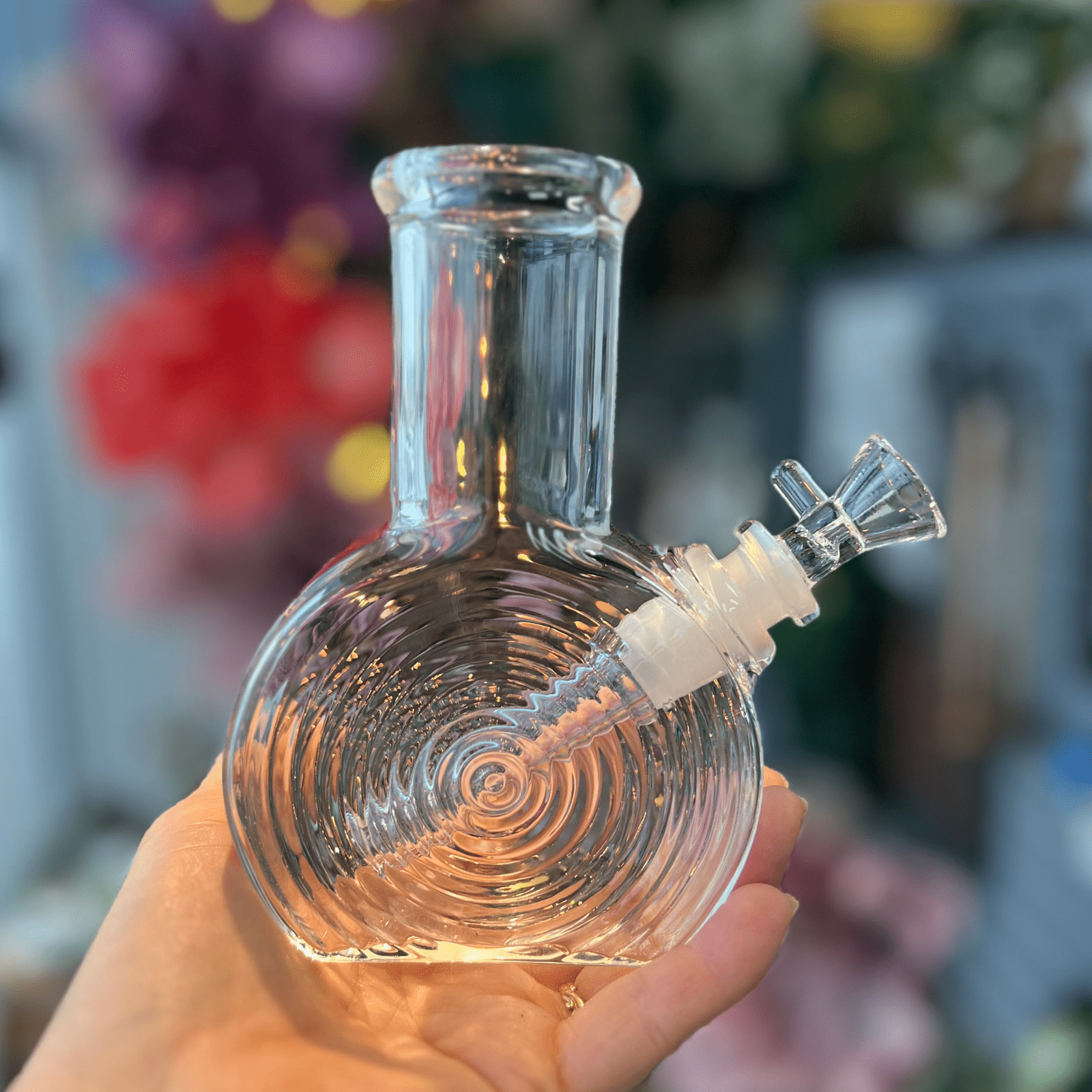 A hand holds “The Art of Smoke Spiral Bong”, a clear glass water pipe with a spiraling design, against a blurred colorful backdrop.