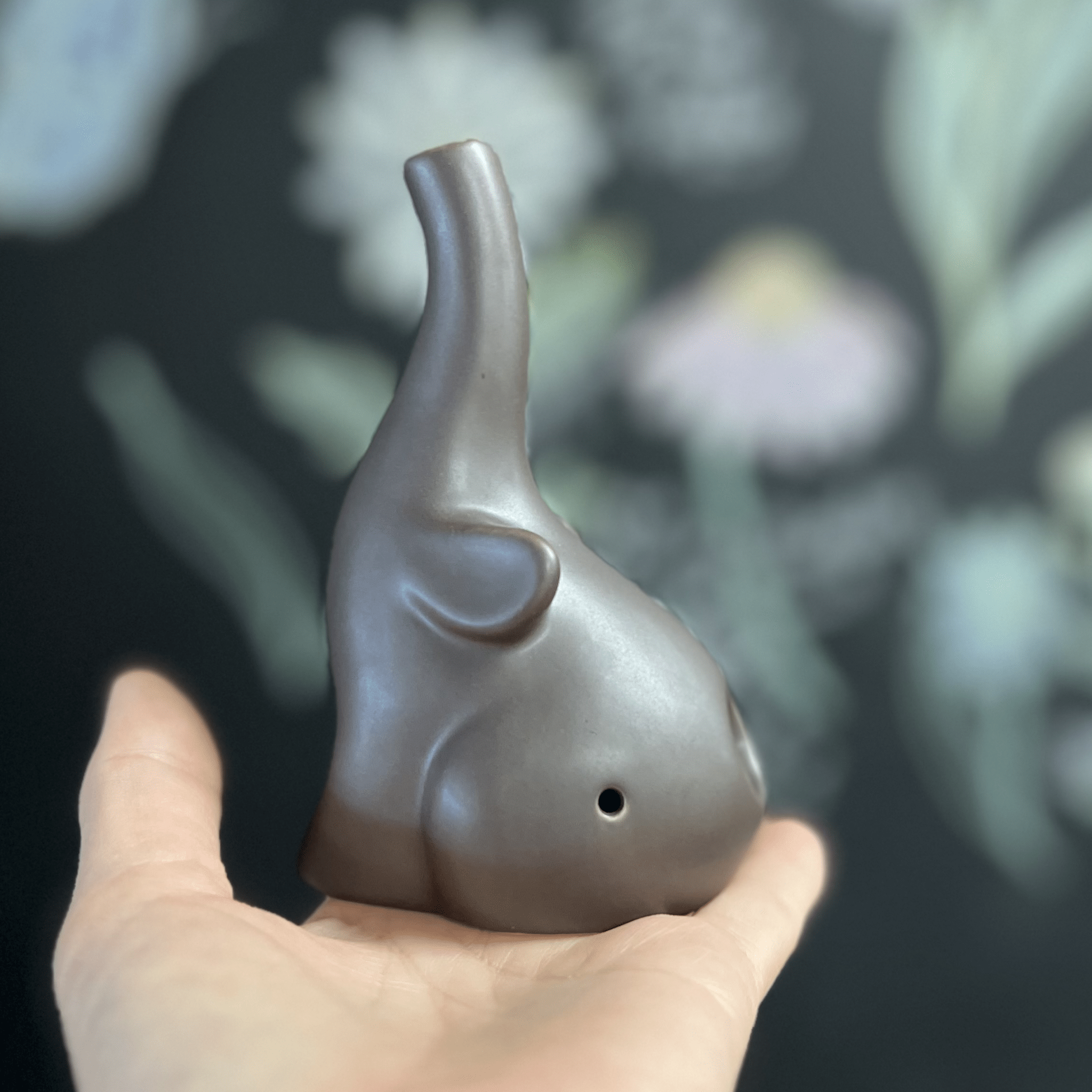 A hand holds the small, gray Baby Elephant Sherlock Pipe, showcasing its unique sculpted form against a blurred green backdrop.