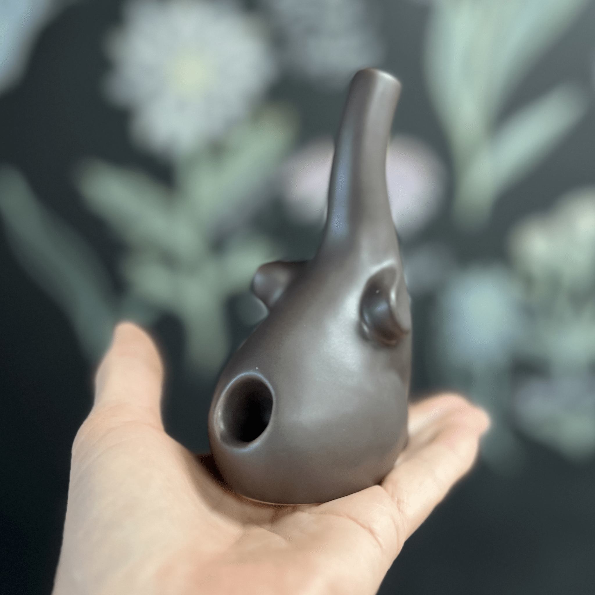 A hand holds the small, dark grey Baby Elephant Sherlock Pipe, angled against a blurred, leafy green background.