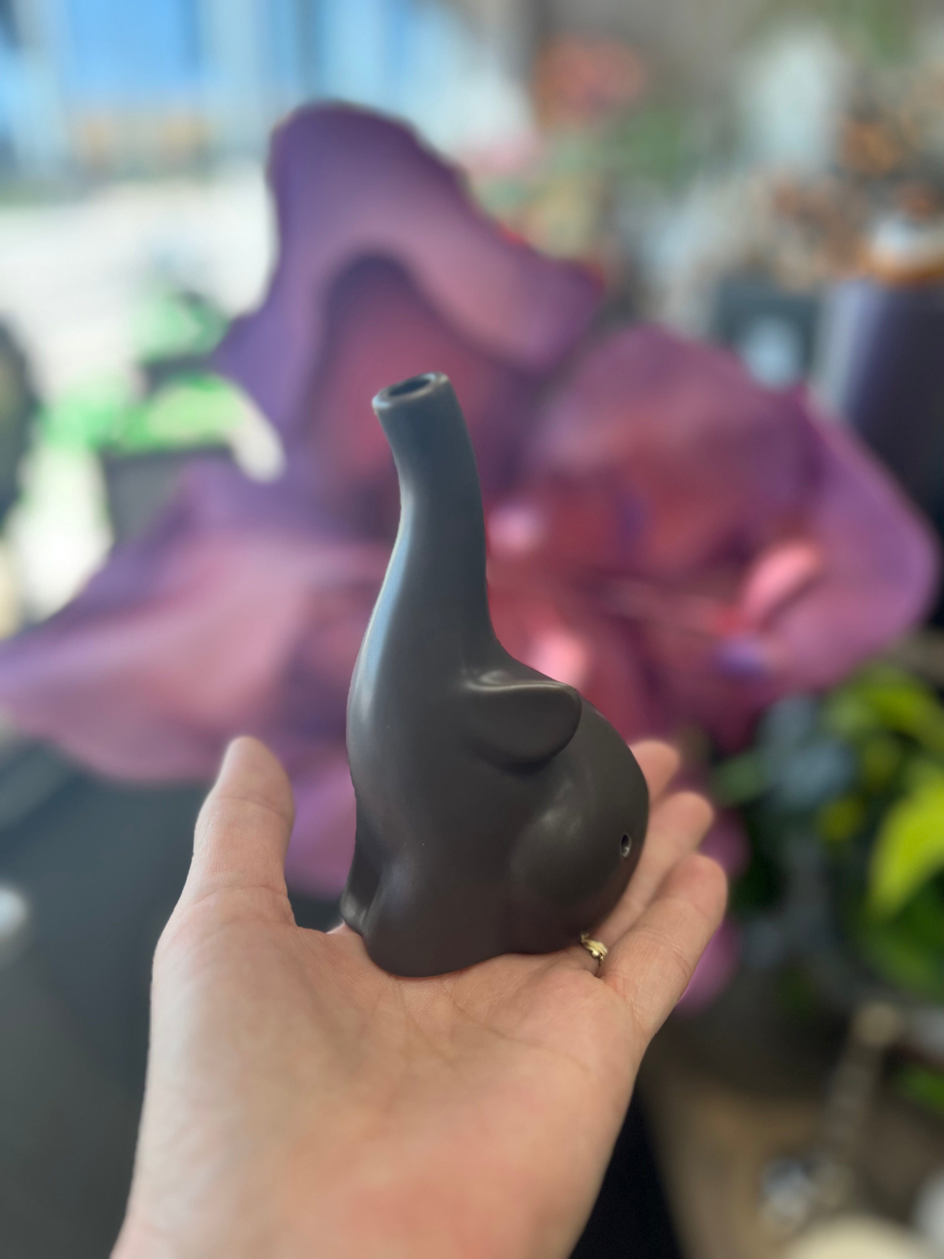 A hand holds the dark-colored Baby Elephant Sherlock Pipe against a vibrant purple backdrop, showcasing its whimsical design.