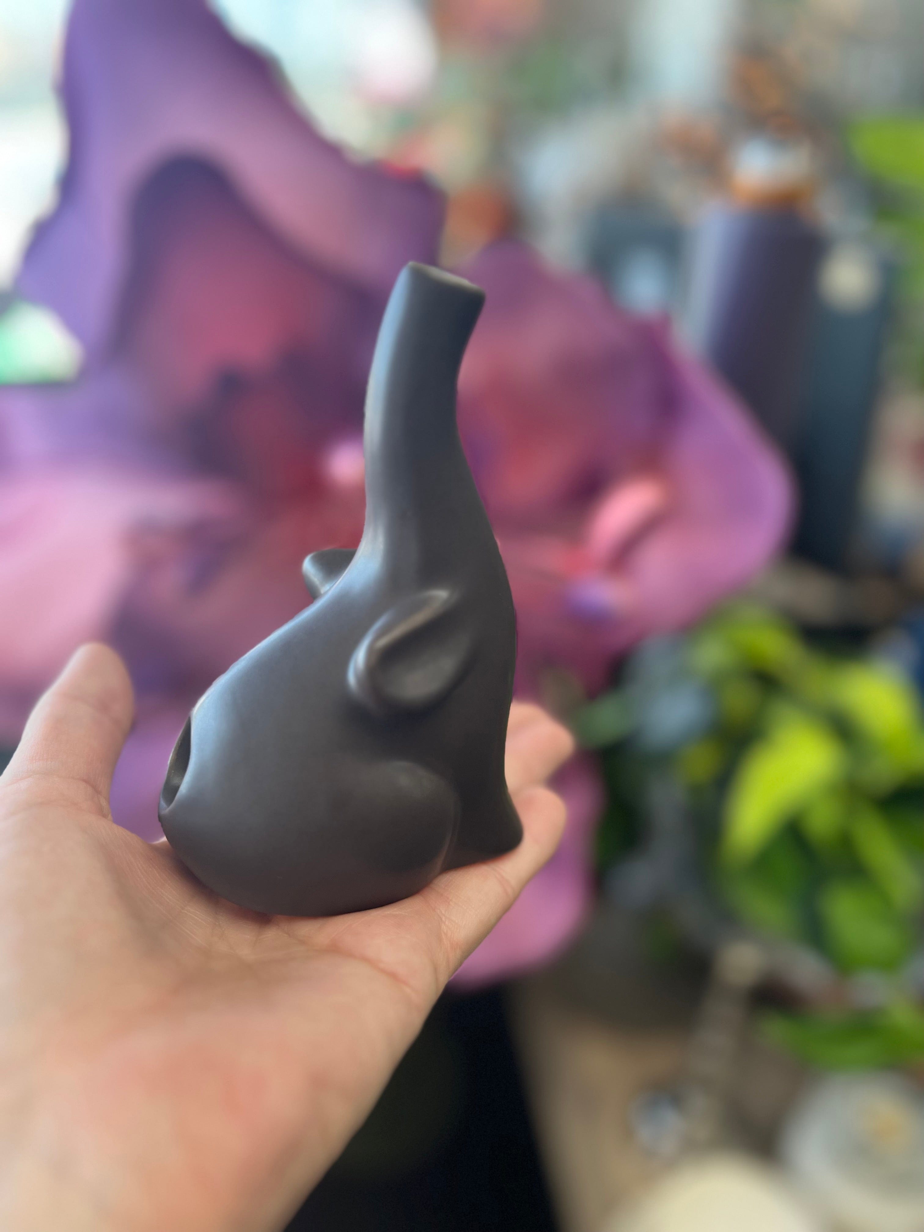 A hand holds the small, black Baby Elephant Sherlock Pipe against a blurred, purple floral backdrop.