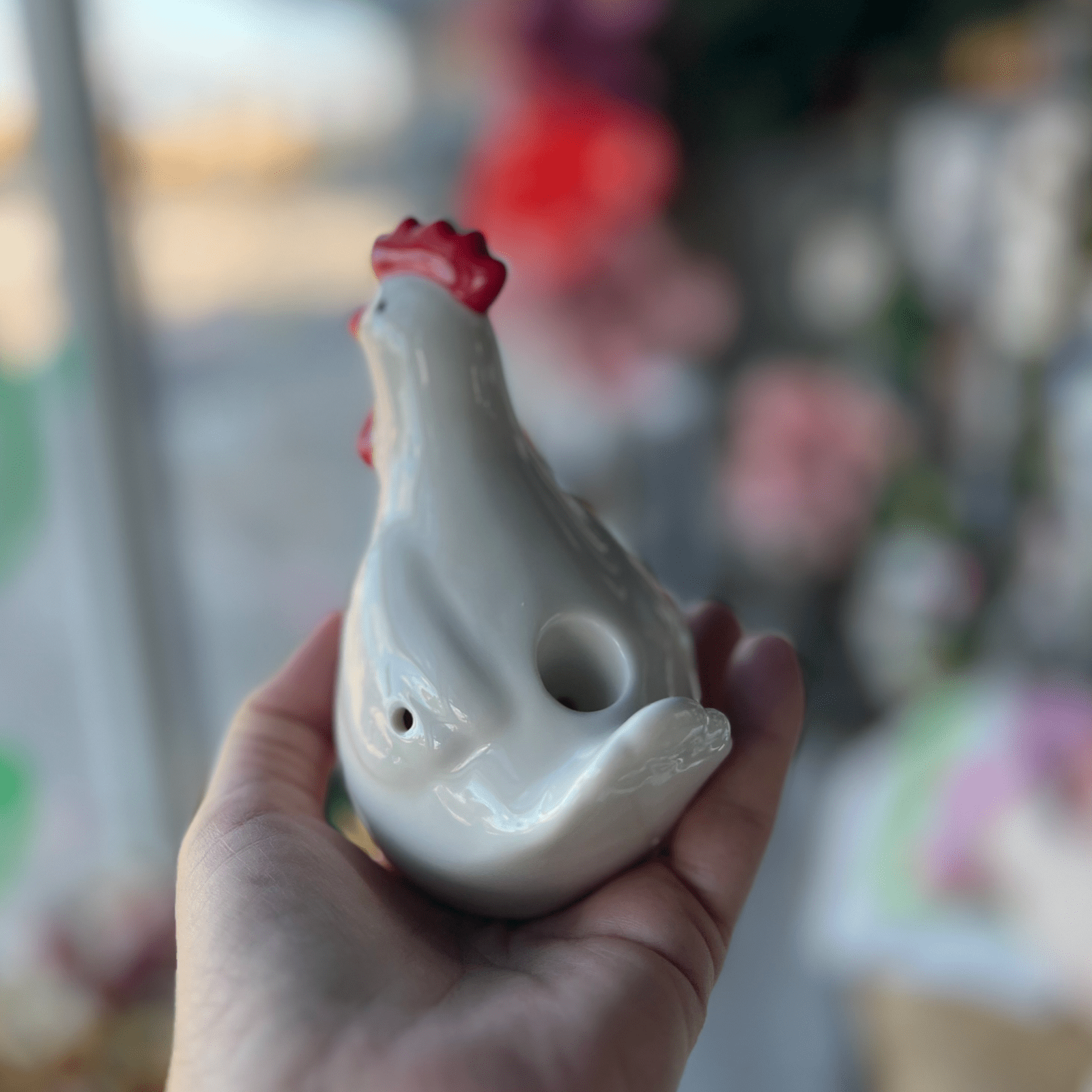 A hand holds the white ceramic Chicken Pipe, shown at a slight angle against a blurred floral background.