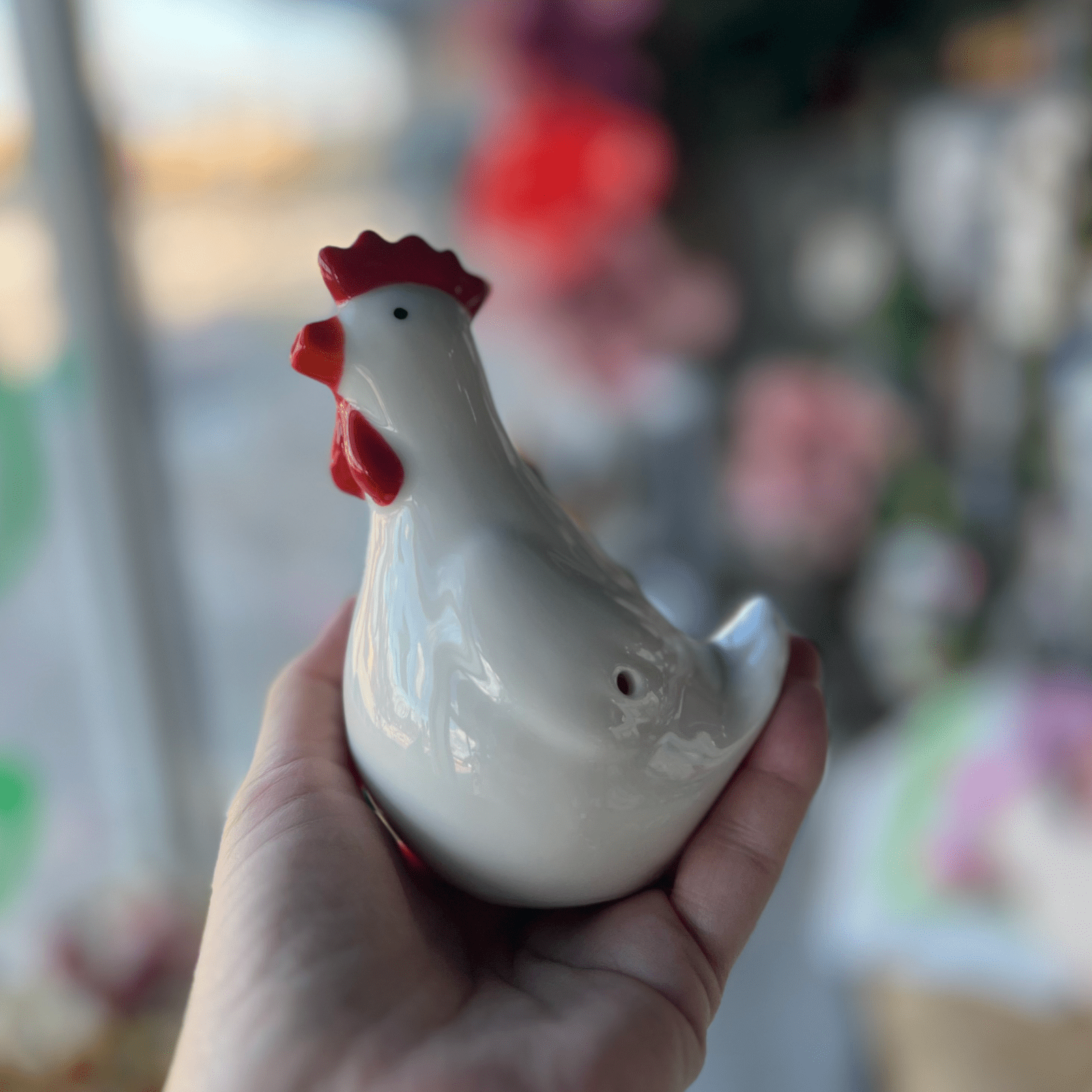 A hand holds the Chicken Pipe, showcasing its smooth, white ceramic finish against a blurred, colorful backdrop.