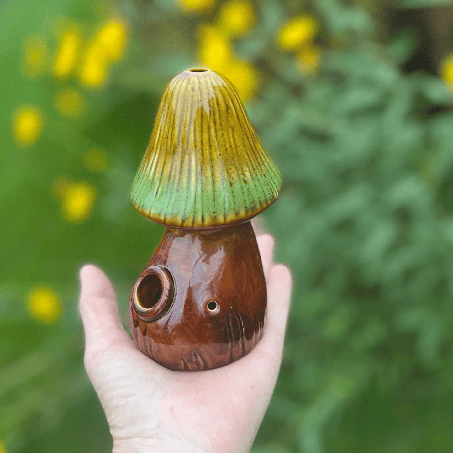 A hand holds the Mushroom Pipe, a green and brown ceramic piece, against a blurred green background.