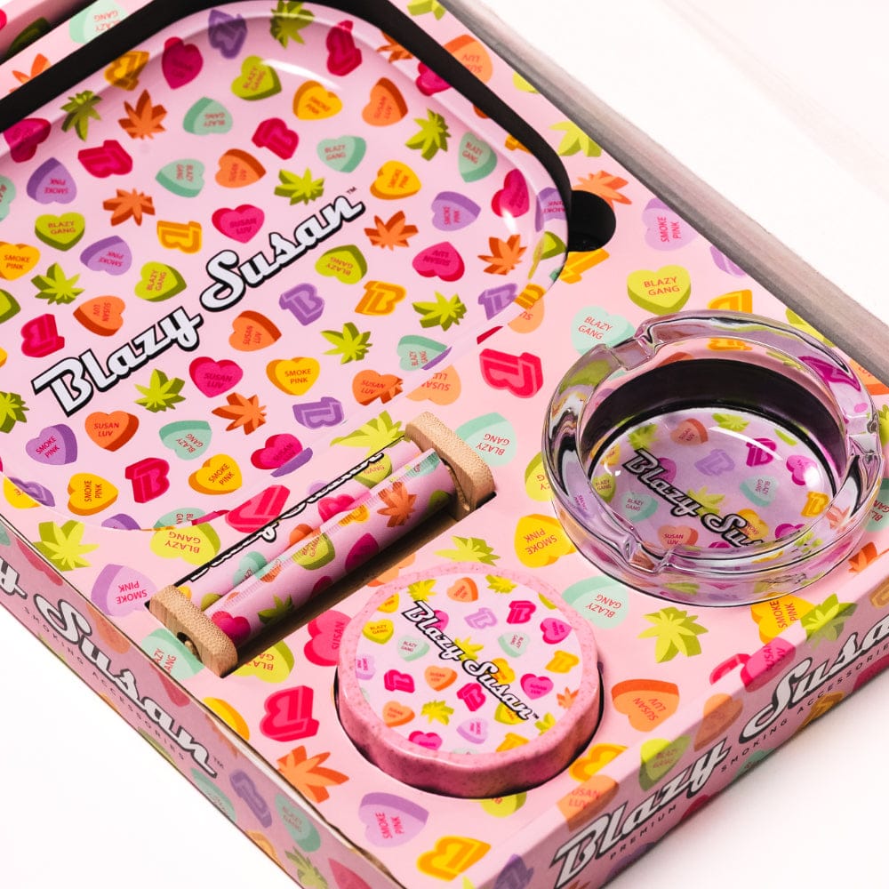 Top-down view of the Blazy Smoker's Kit showing a pink patterned tray, glass ashtray, rolling wheel, and wooden-handled rolling paper holder.