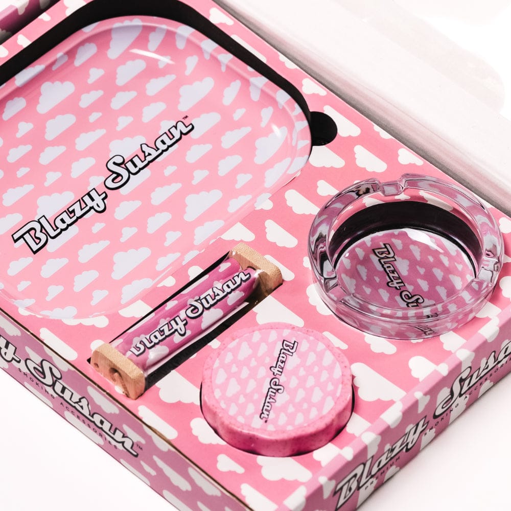 Angled close-up of the pink cloud–patterned Blazy Smoker's Kit showing a tray, glass ashtray, roller and round grinder nestled in branded packaging.