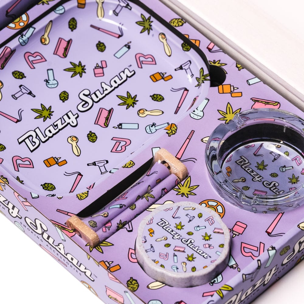 Top-down view of a purple Blazy Smoker's Kit tray and accessories patterned with small cannabis leaves, pipes, lighters, and a clear glass ashtray.