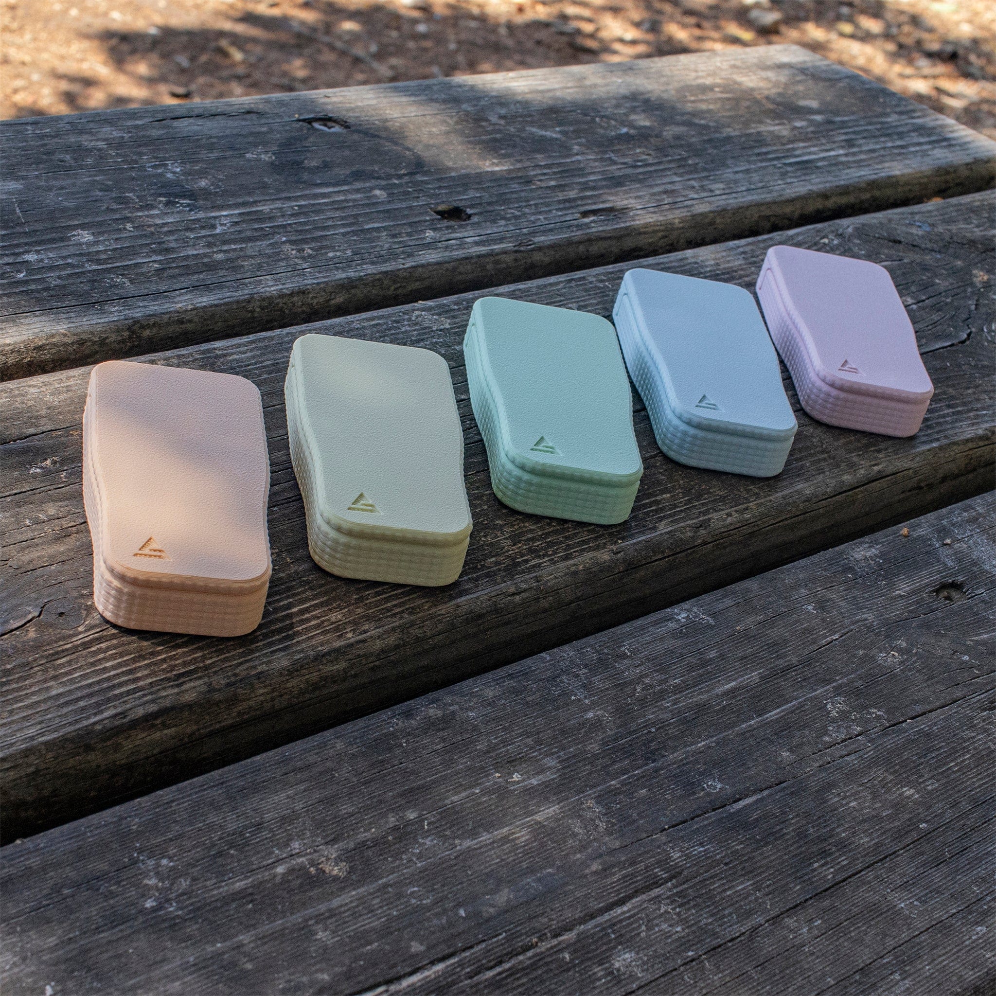 Angled close-up of several STASURE Joint Case Pastel Polar Blue cases in soft pastel tones lined up on a weathered wooden picnic table