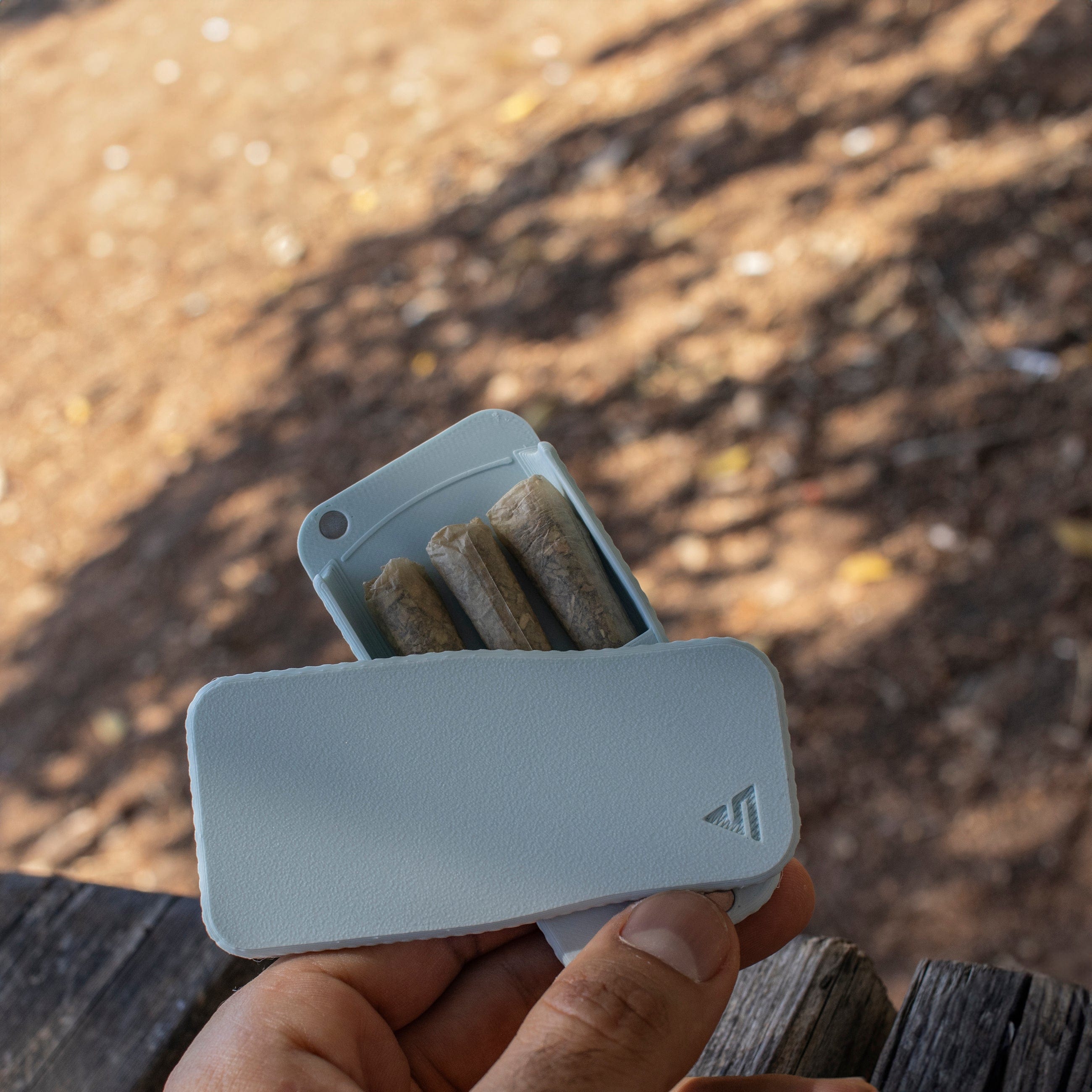 Handheld, angled close-up of the STASURE Joint Case Pastel Polar Blue opened to show rolled papers inside against a blurred outdoor background