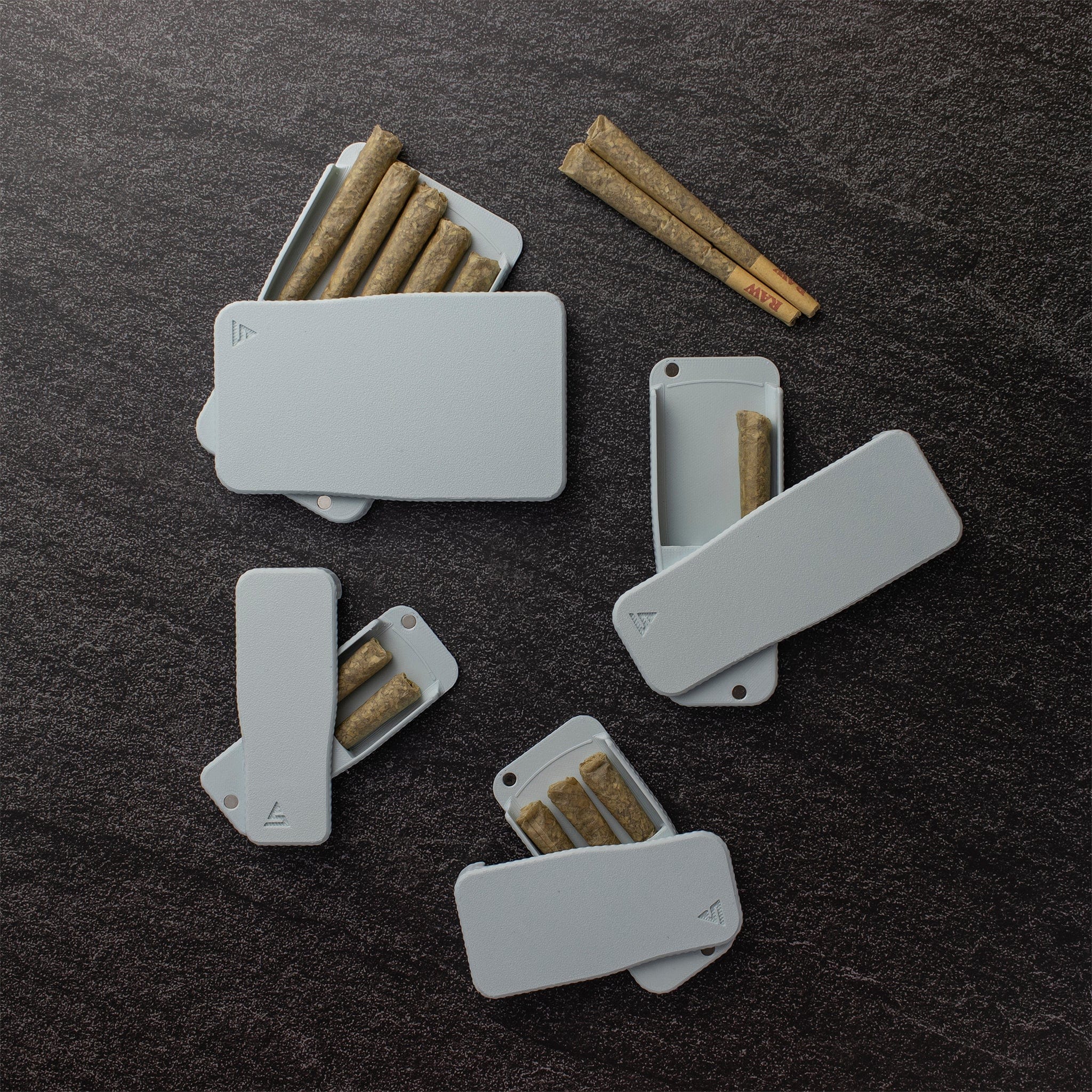 Top-down view of multiple STASURE Joint Case Pastel Polar Blue cases open on a dark textured surface, each holding rolled joints.