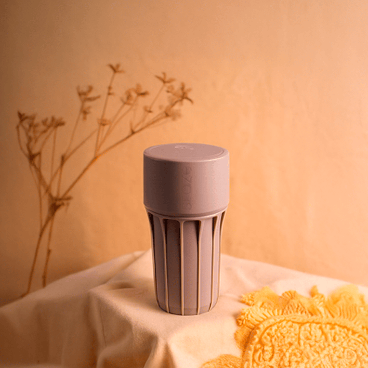 Center-frontal view of the Foam Staze Preserve cylinder standing on draped fabric with soft warm lighting and dried stems blurred in the background