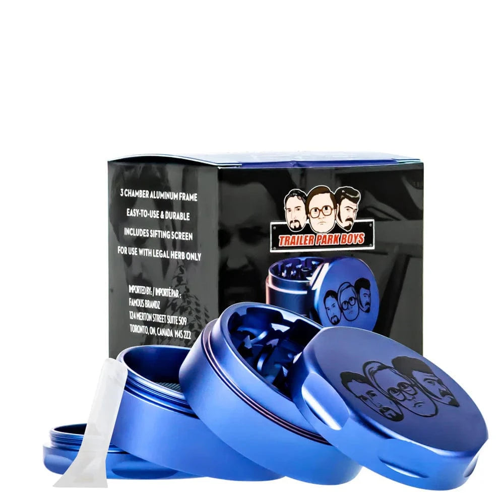 Angled close-up of the blue Famous X Trailer Park Boys Grinder - Assorted Colors with its three open aluminum chambers, embossed lid art, and packaging behind.