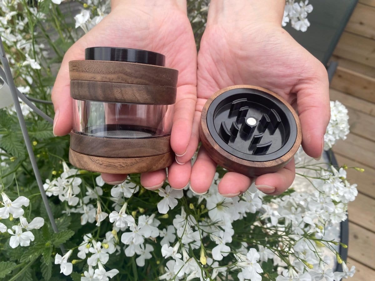 A close-up, overhead view showcases The Bzz 4 Piece Grinder held in two hands against a backdrop of white flowers.