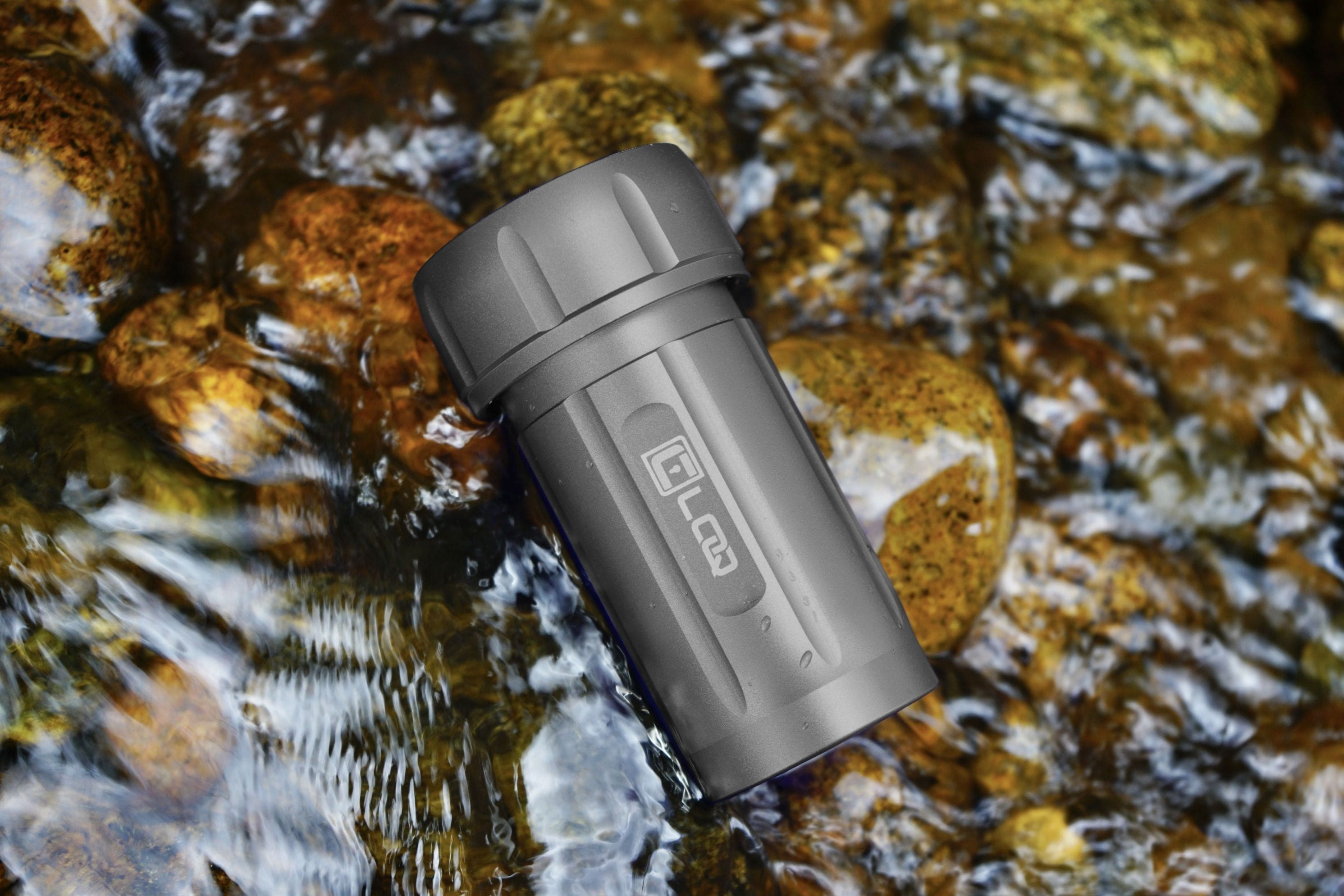 The Zytel Gray Premium 120CC Storage Canister rests against smooth river rocks, captured in a low-angle, natural light shot.