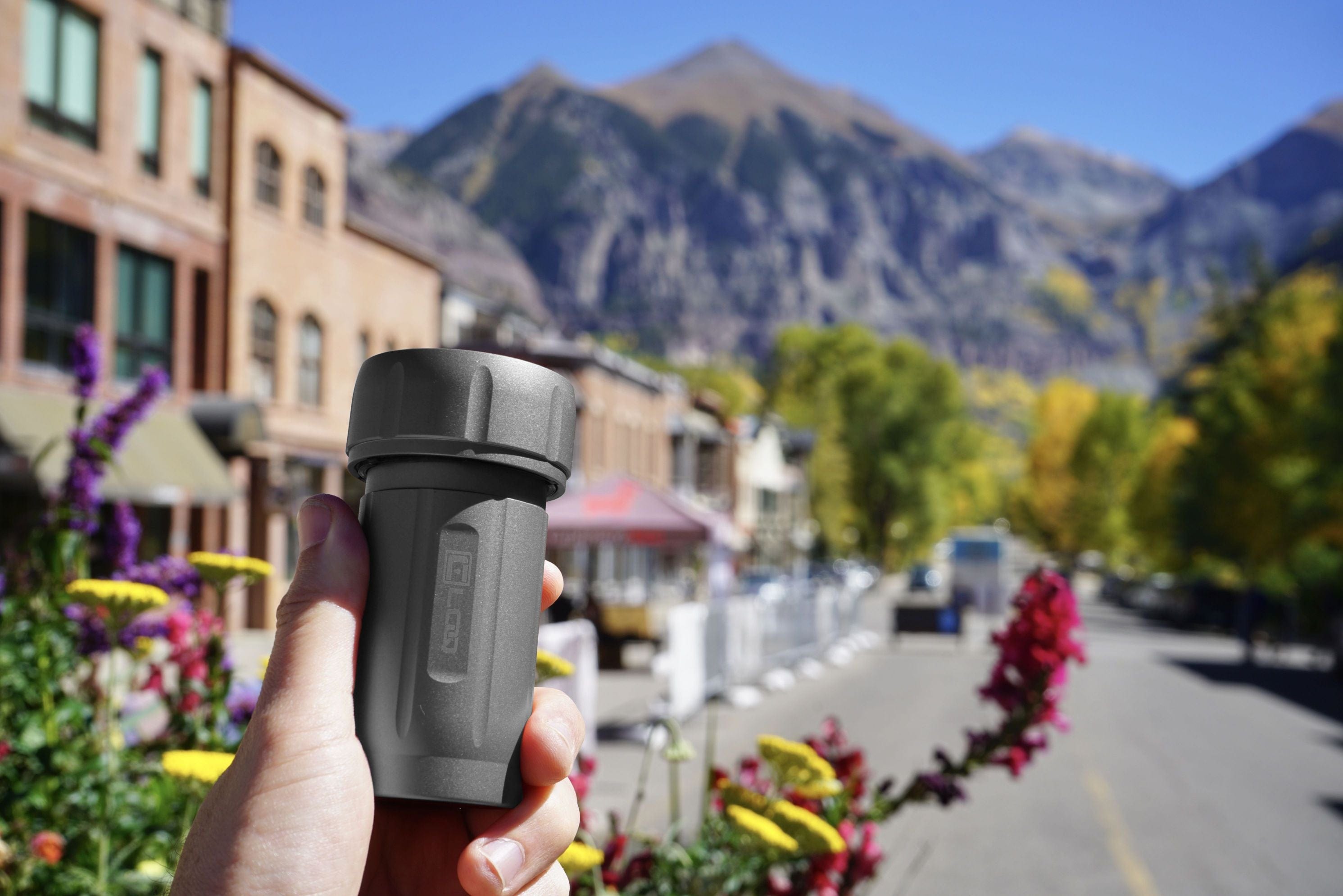 A hand holds the Zytel Gray Premium 120CC Storage Canister with a black cap, framed by colorful flowers and a mountain backdrop.