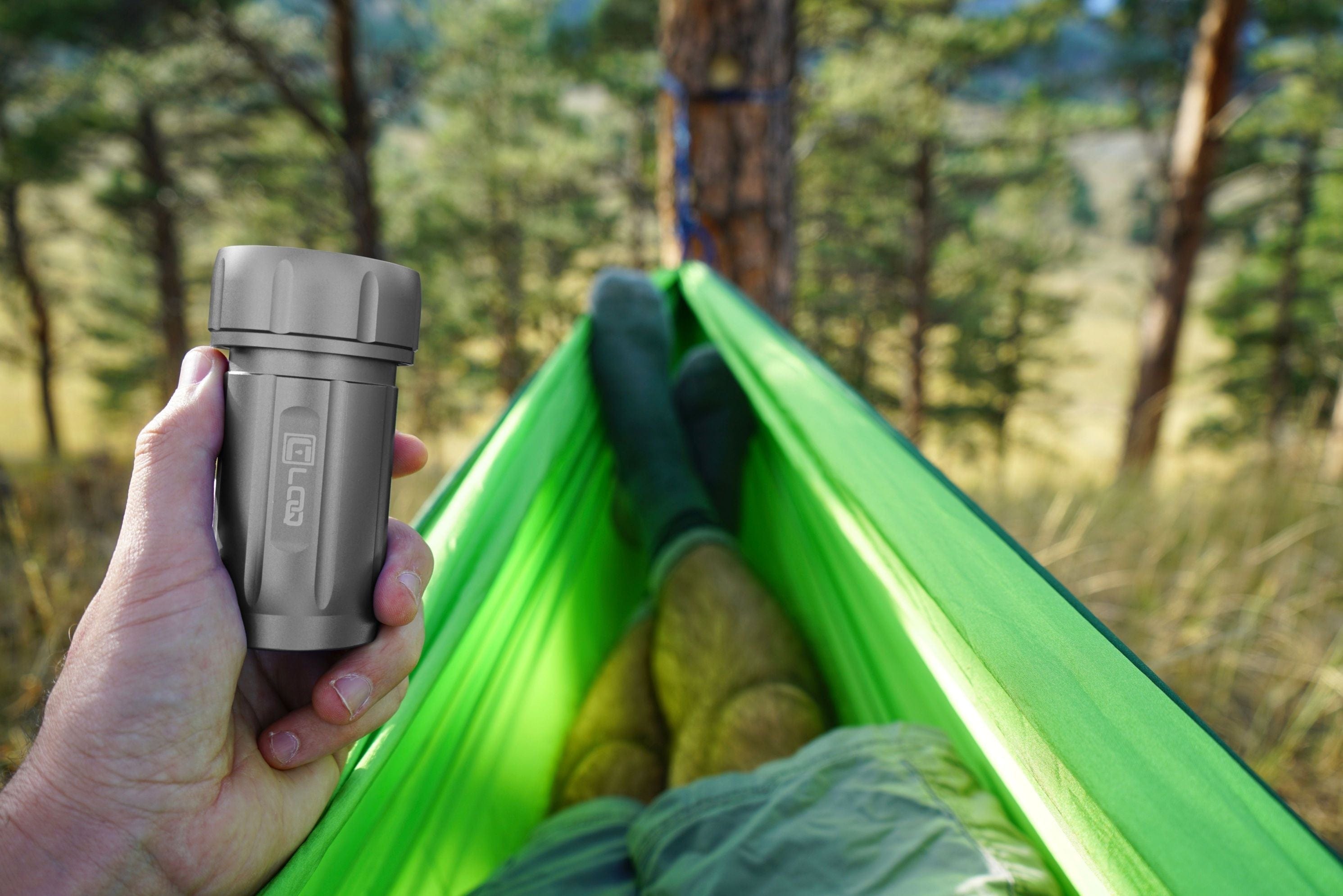Handheld close-up of the Premium 120CC Storage Canister | Zytel Gray | Black Cap in a left hand with a blurred green hammock and forest background.