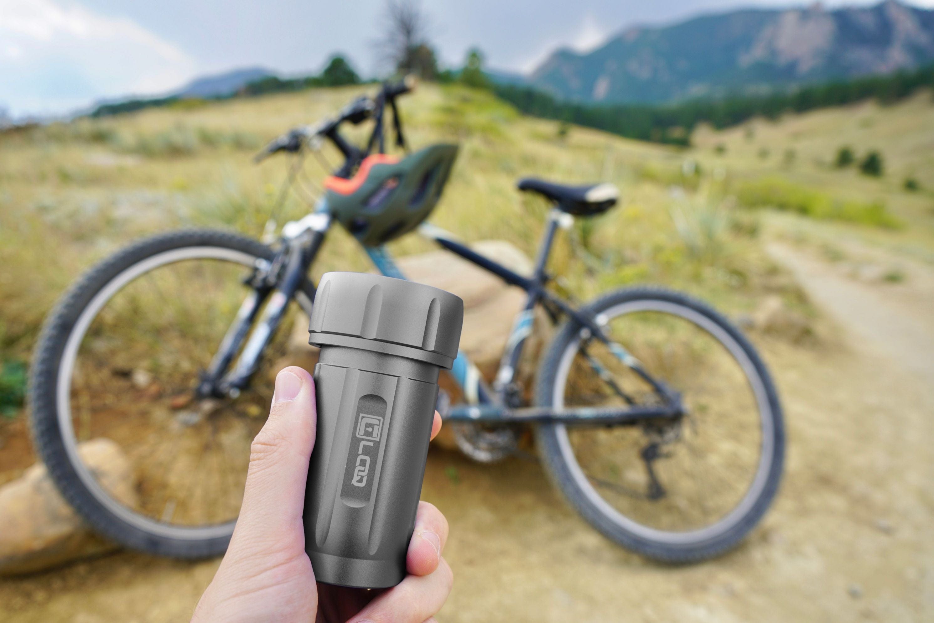 A hand holds the Zytel Gray Premium 120CC Storage Canister against a blurred background of a bicycle and distant hills.
