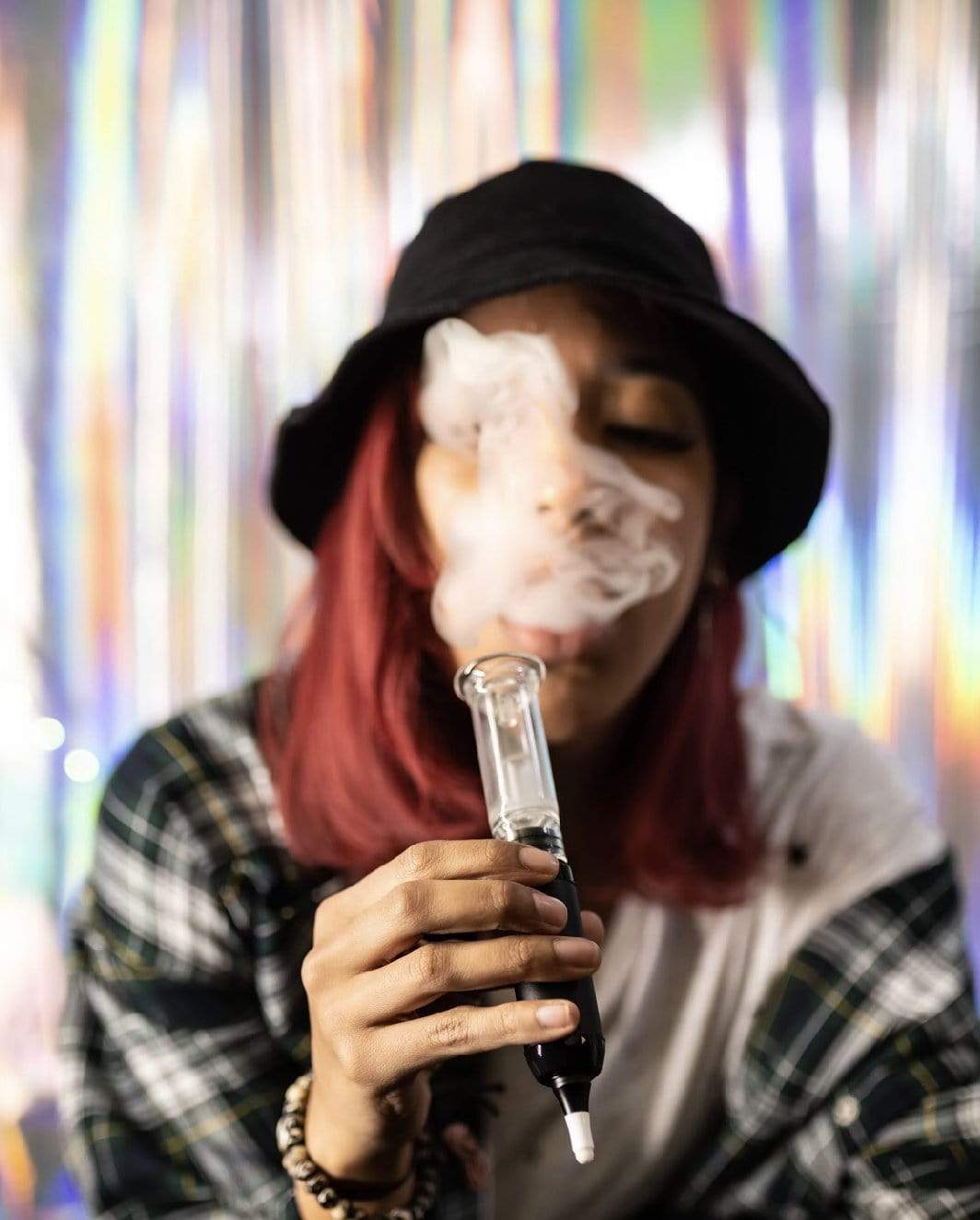 A close-up, front view shows smoke rising from the DabTech Trio as it’s held against a blurred, colorful backdrop.