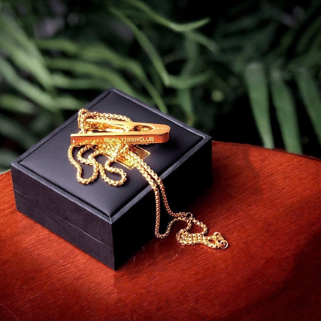 A close-up shot of the gold Daily High Club Roach Clip displayed on a black velvet surface.