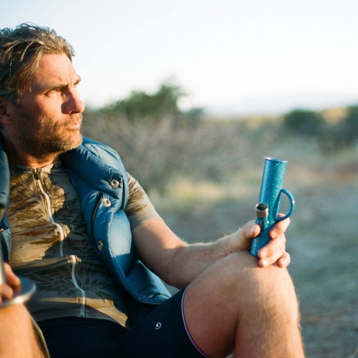 A man holds the DangleBong™️ Titanium Water Pipe in a field, with a low angle emphasizing its sleek, metallic form against the distant landscape.