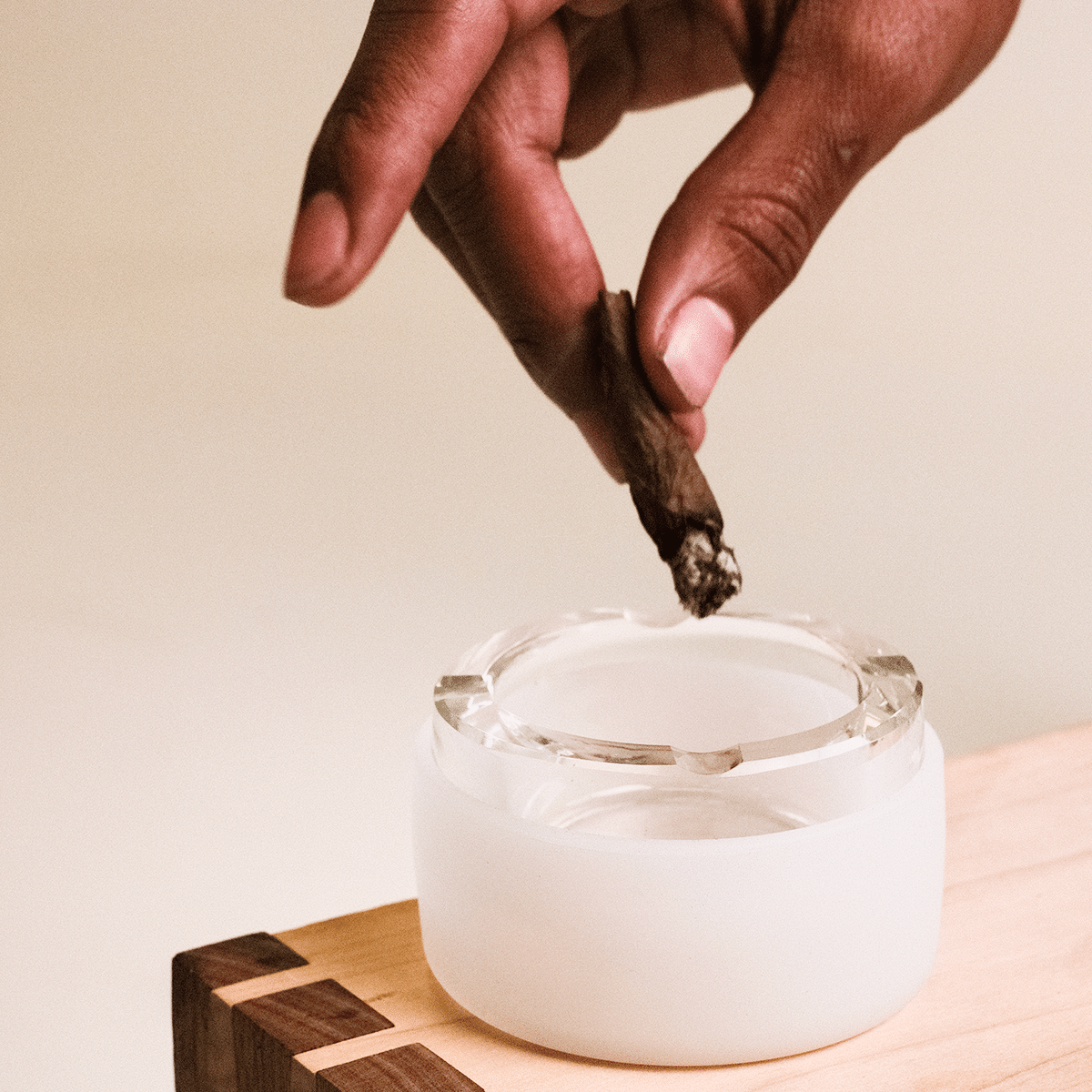 A hand holds a lit cigarette above the Ashtray, viewed from a slightly elevated angle against a neutral backdrop.