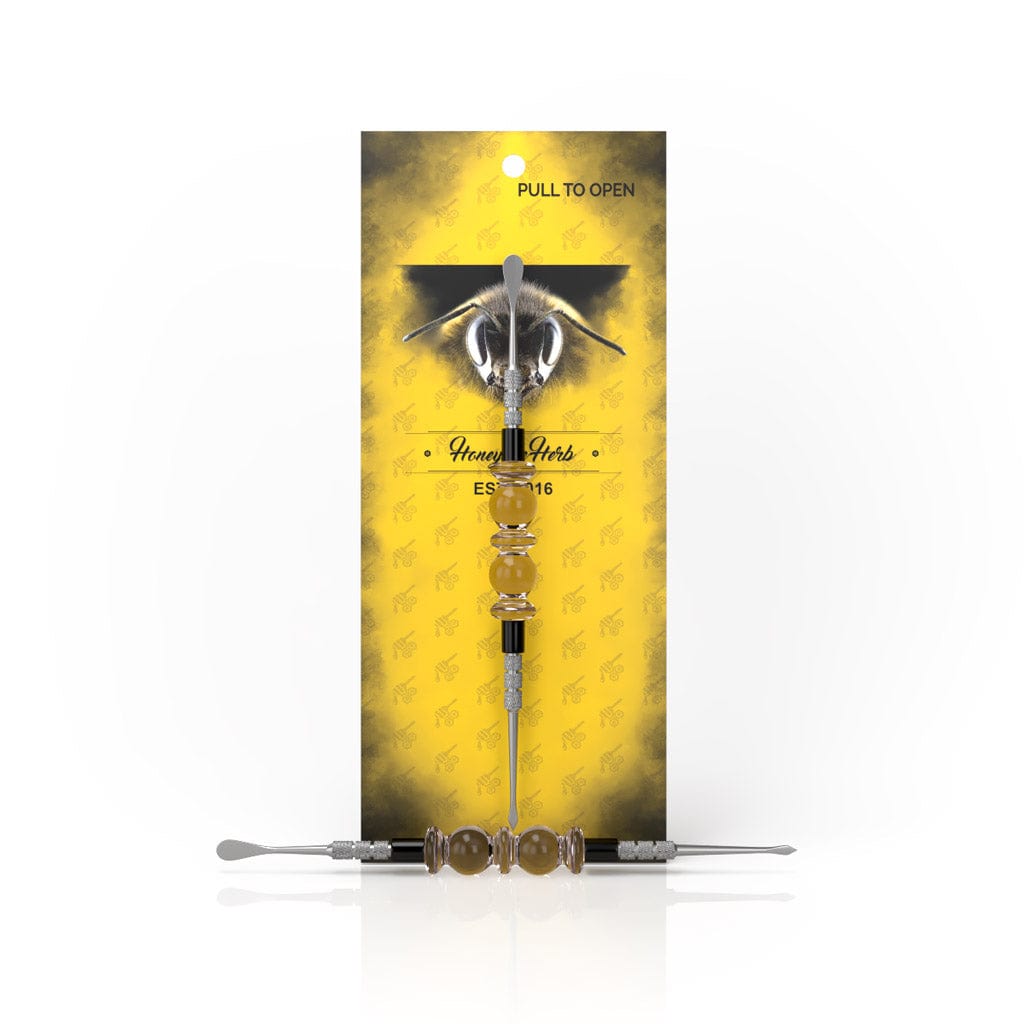 Front view of the Bliss Dab Tool, a silver and gold metallic tool set against a dark yellow background.