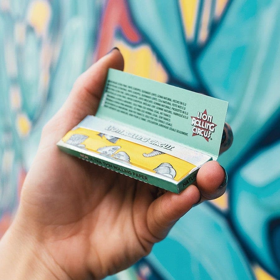 A hand holds a pack of Lion Rolling Circus Silver Line Ultra Thin 1-1/4 Rolling Papers against a blurred, colorful backdrop.