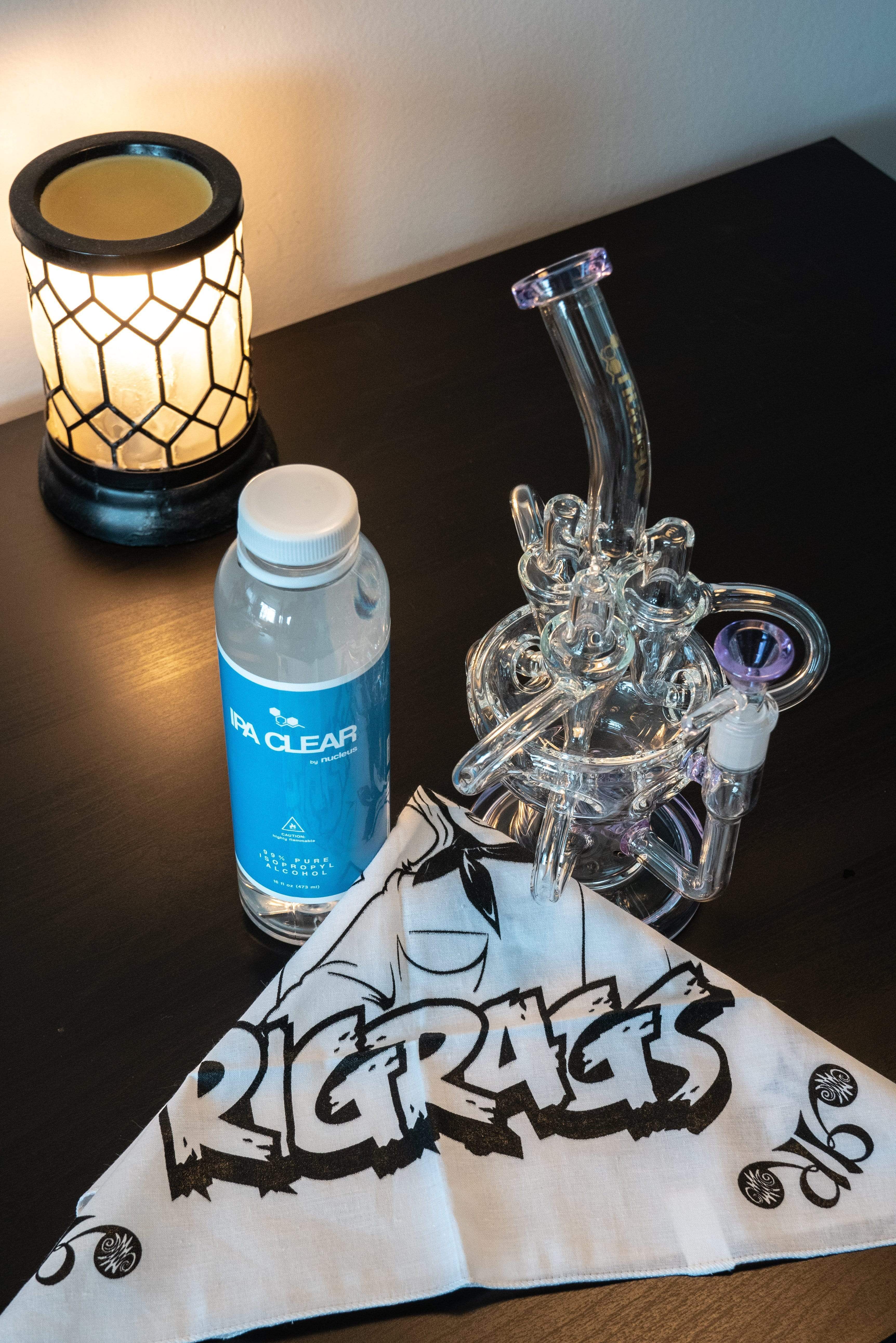 A close-up, angled view shows the bottle of Pure Isopropyl Alcohol next to a clear glass pipe on a dark surface.