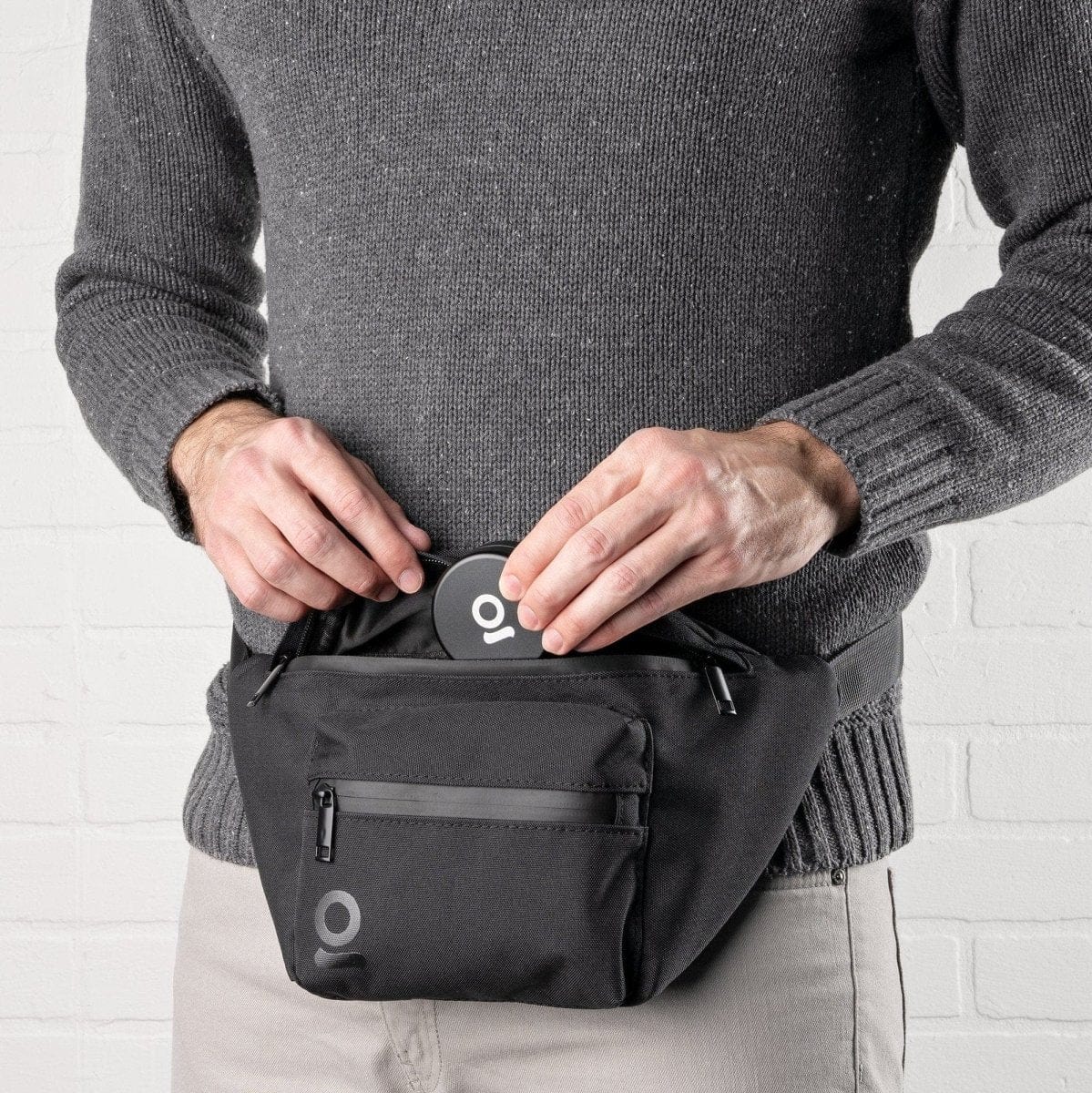 A close-up, angled view shows the Carbon-lined Fanny Pack / Travel Pouch being zipped by a hand, highlighting its compact size and dark color.