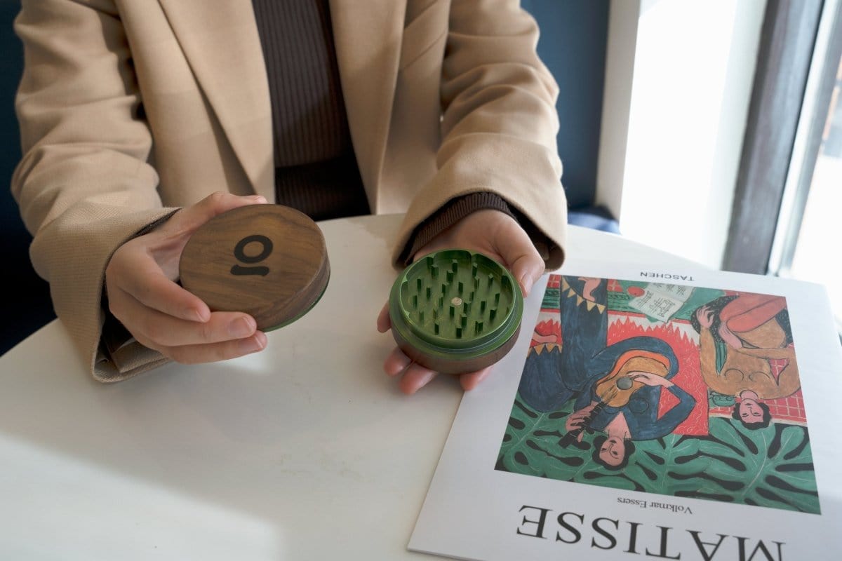 A close-up, angled view shows the 3 Inch, 2-Piece Walnut Grinder held in a person's hand against a patterned book cover.
