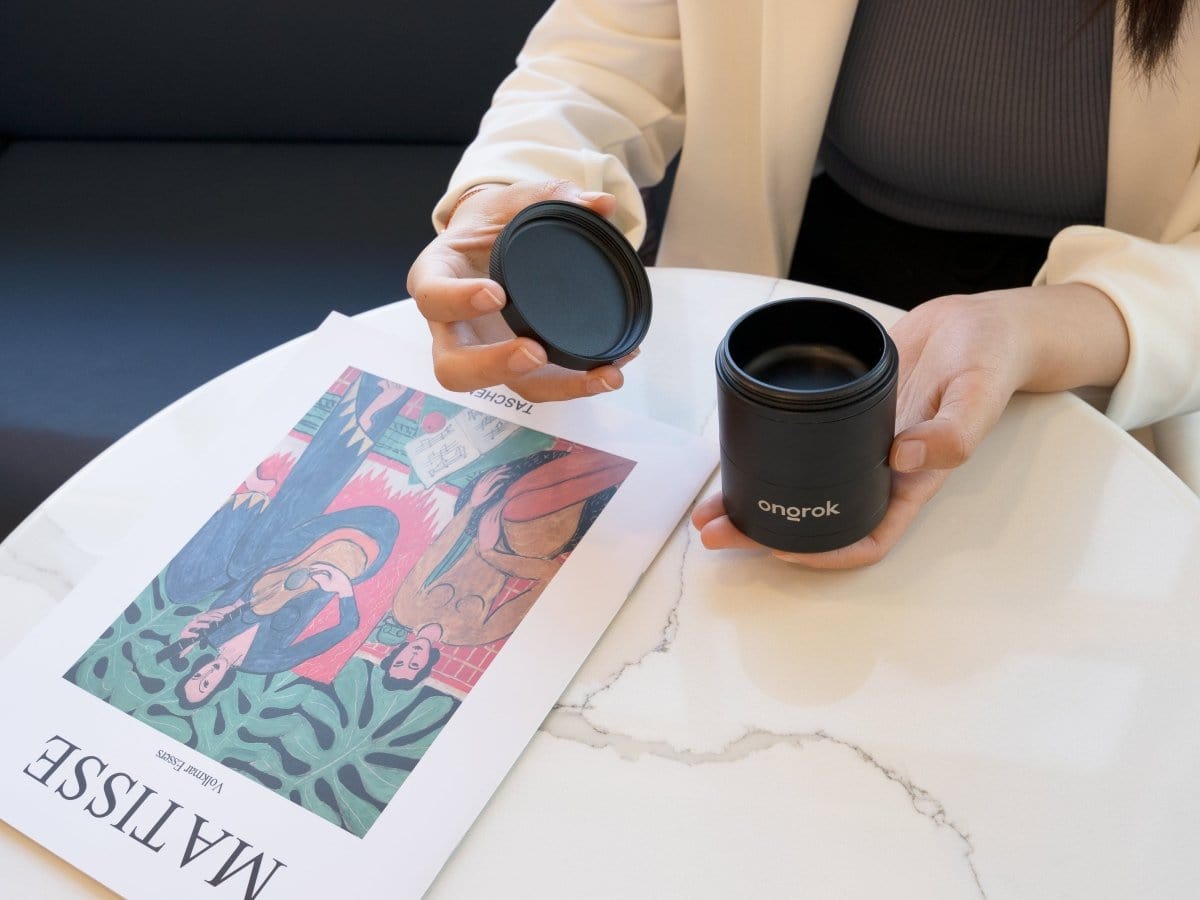 A hand holds the 5 Piece Storage Grinder, set against a blurred background with text and a dark surface.