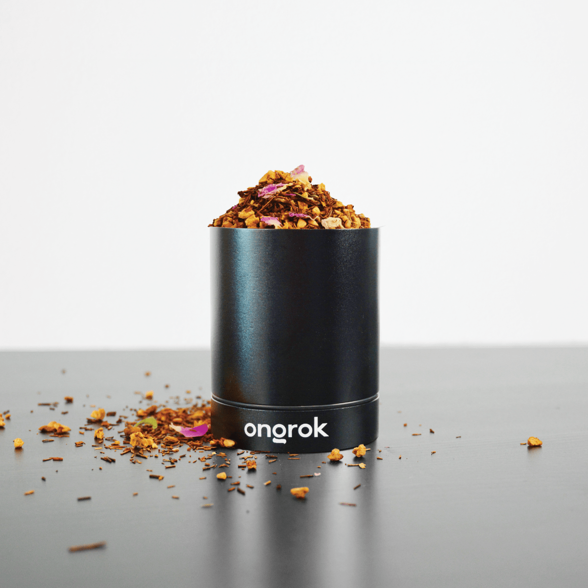 The Aluminum Metal Storage Jar sits on a dark surface surrounded by scattered plant material, captured in a low-angle studio shot.